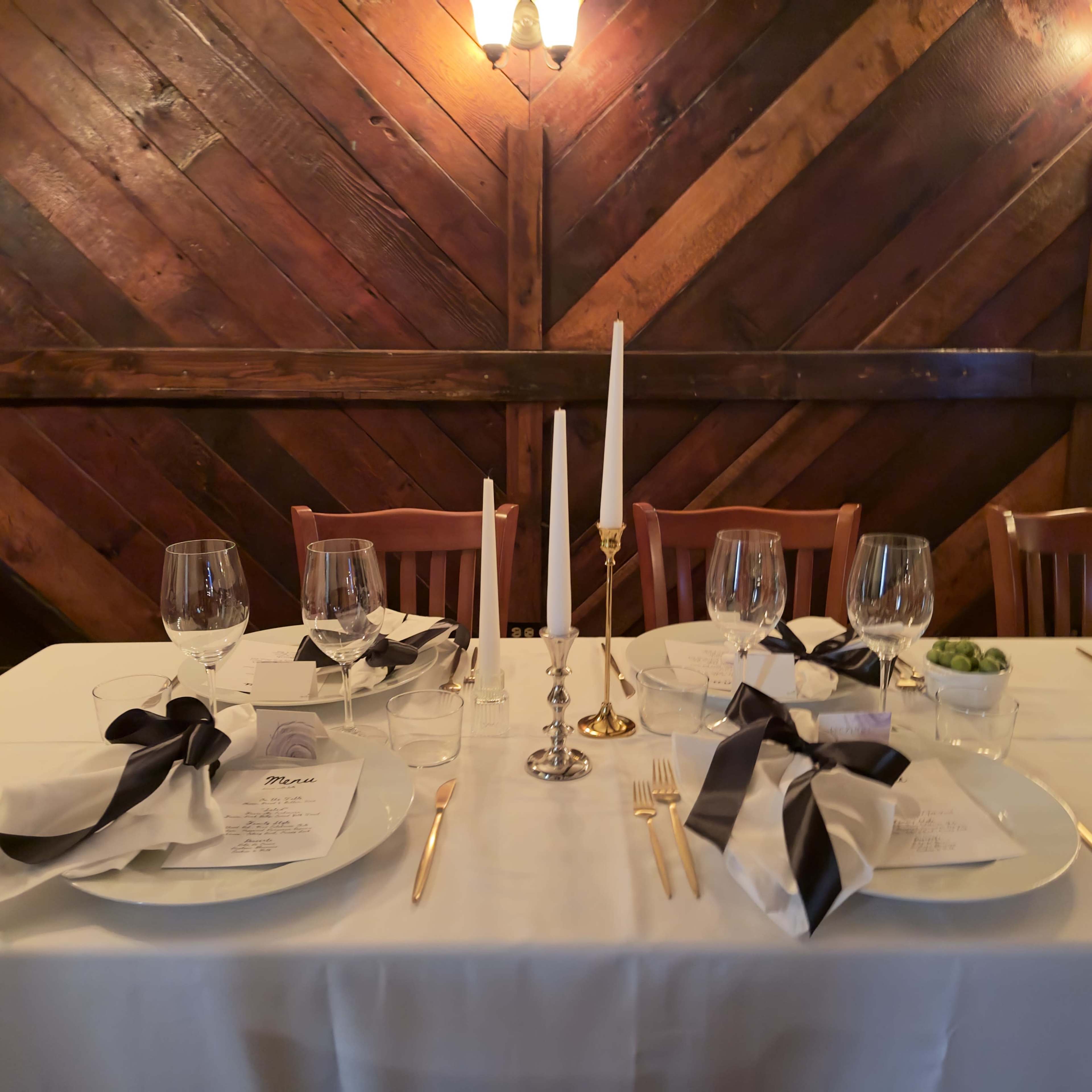 A formal dining table is set with white tablecloths, elegant dishware, glasses, and candles against a wooden wall.