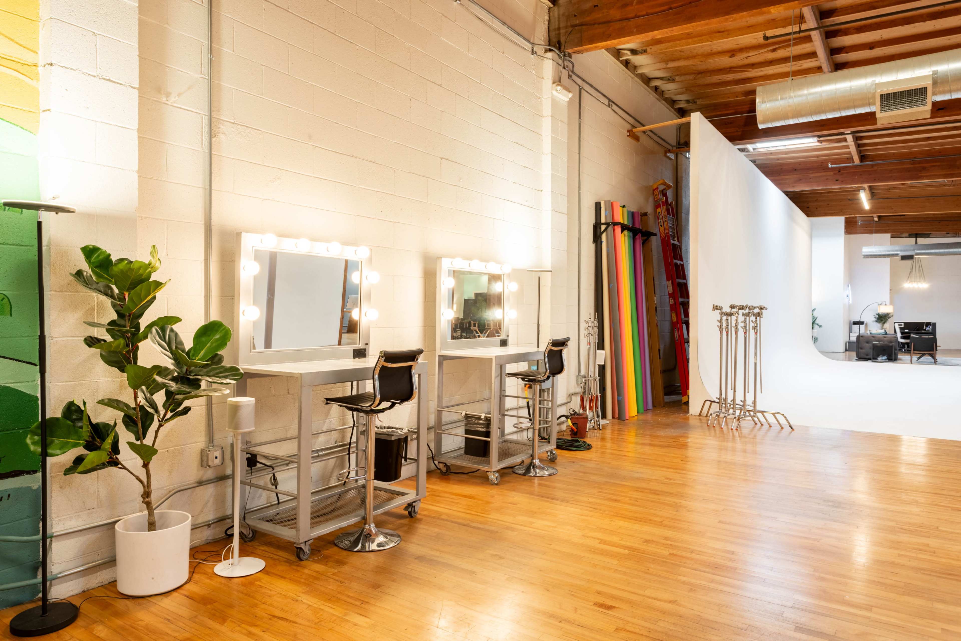 The image shows a spacious salon or photography studio featuring two mirrored vanities with bright bulbs, black stools, and a wooden floor, alongside a white backdrop and colorful panels in the background.