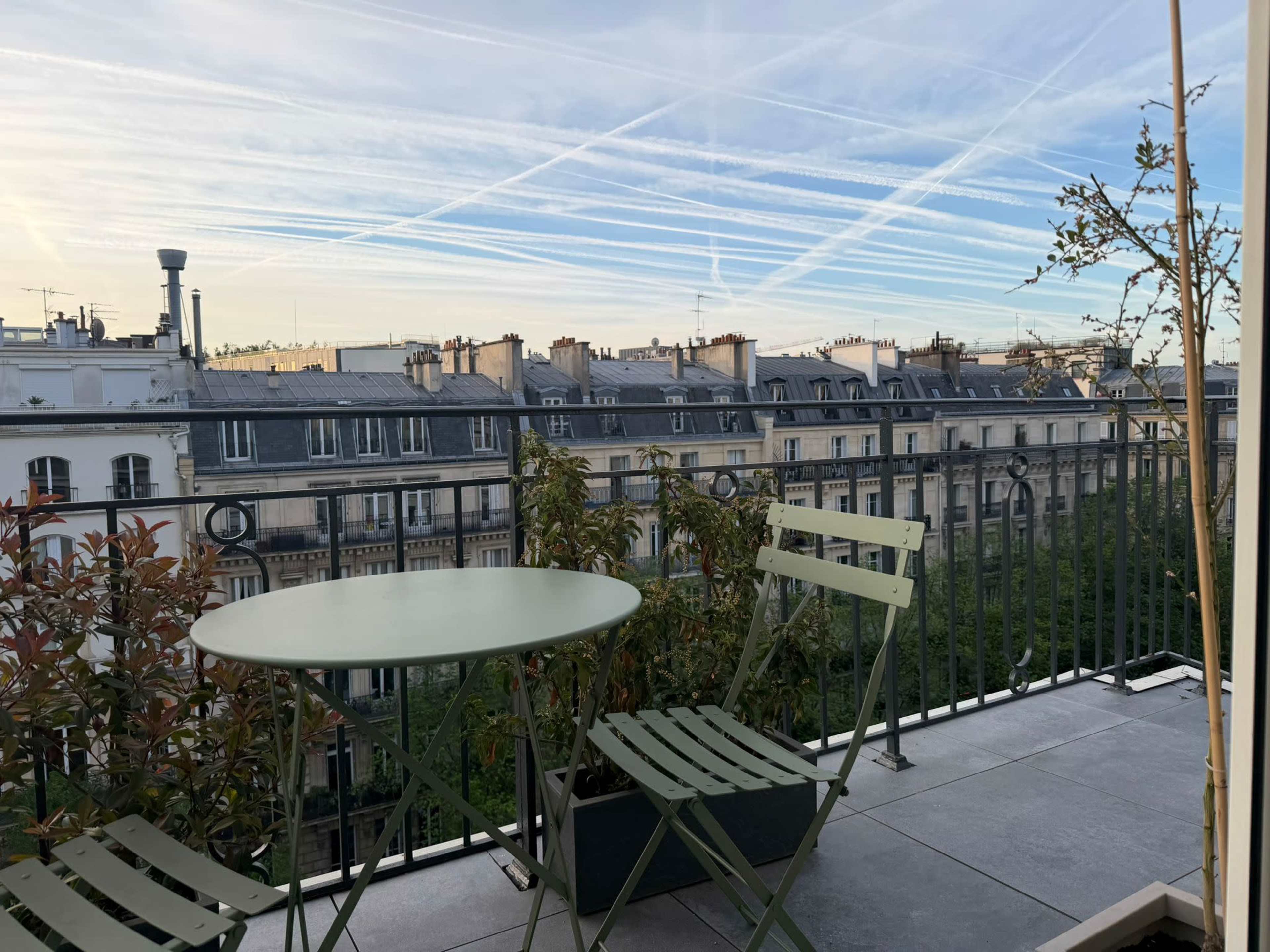 The image shows a small terrace with a green table and two chairs, overlooking Parisian buildings under a sky marked by contrails.