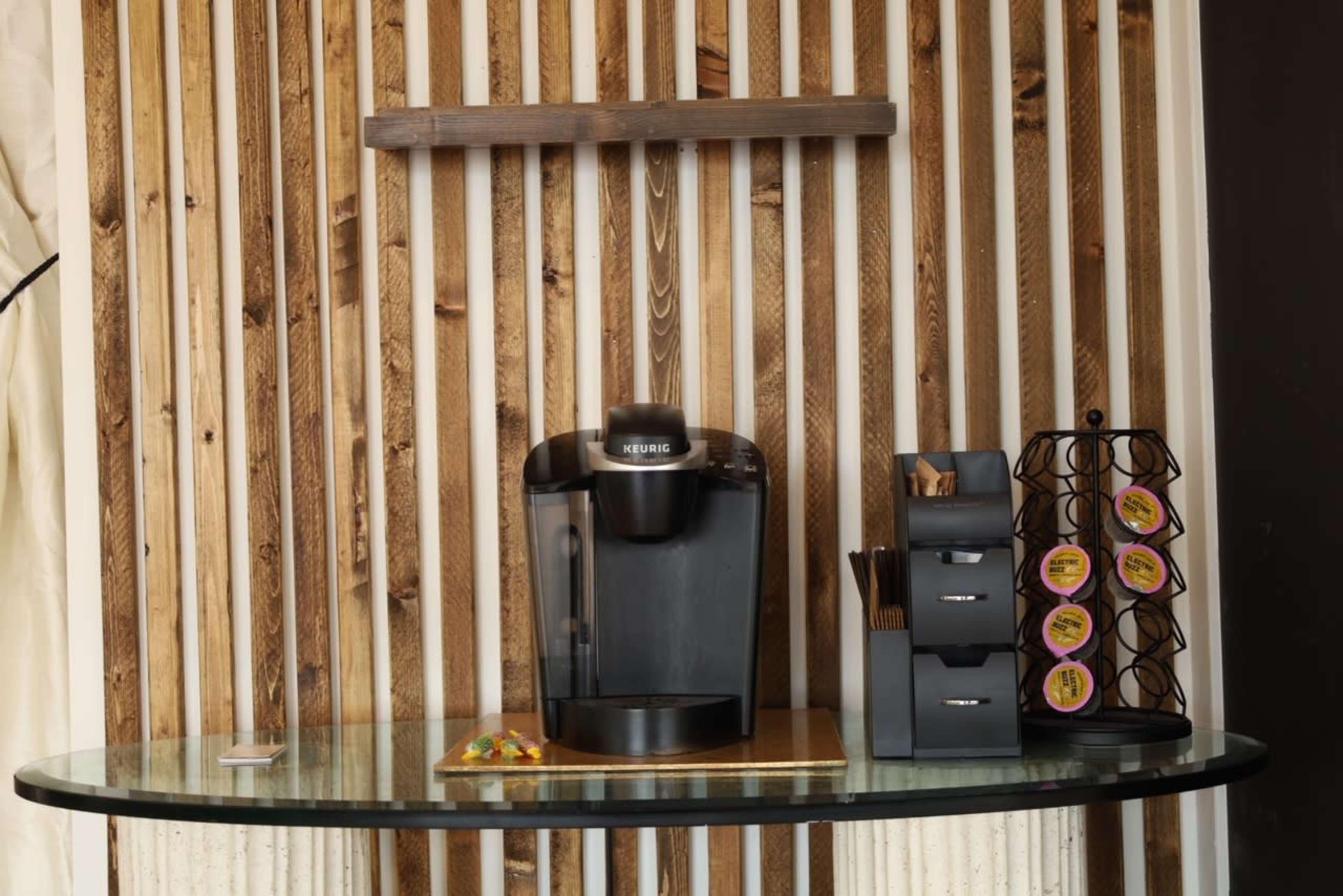 The image shows a coffee station featuring a Keurig machine on a glass table, accompanied by a stand for coffee pods and a container with utensils against a wooden slat backdrop.