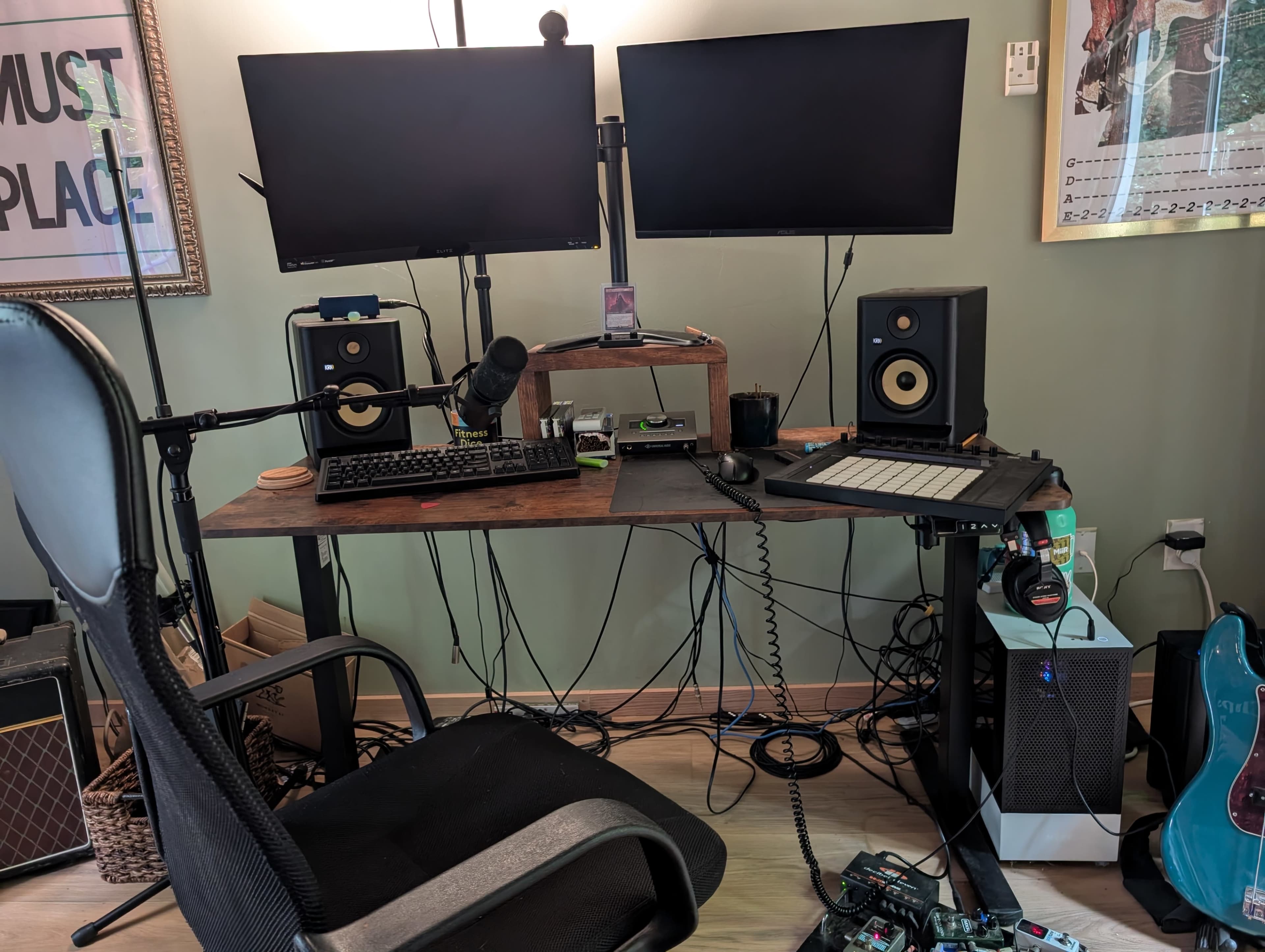 A workspace is set up with two monitors, speakers, a microphone, and various cables on a wooden desk, accompanied by a black office chair.