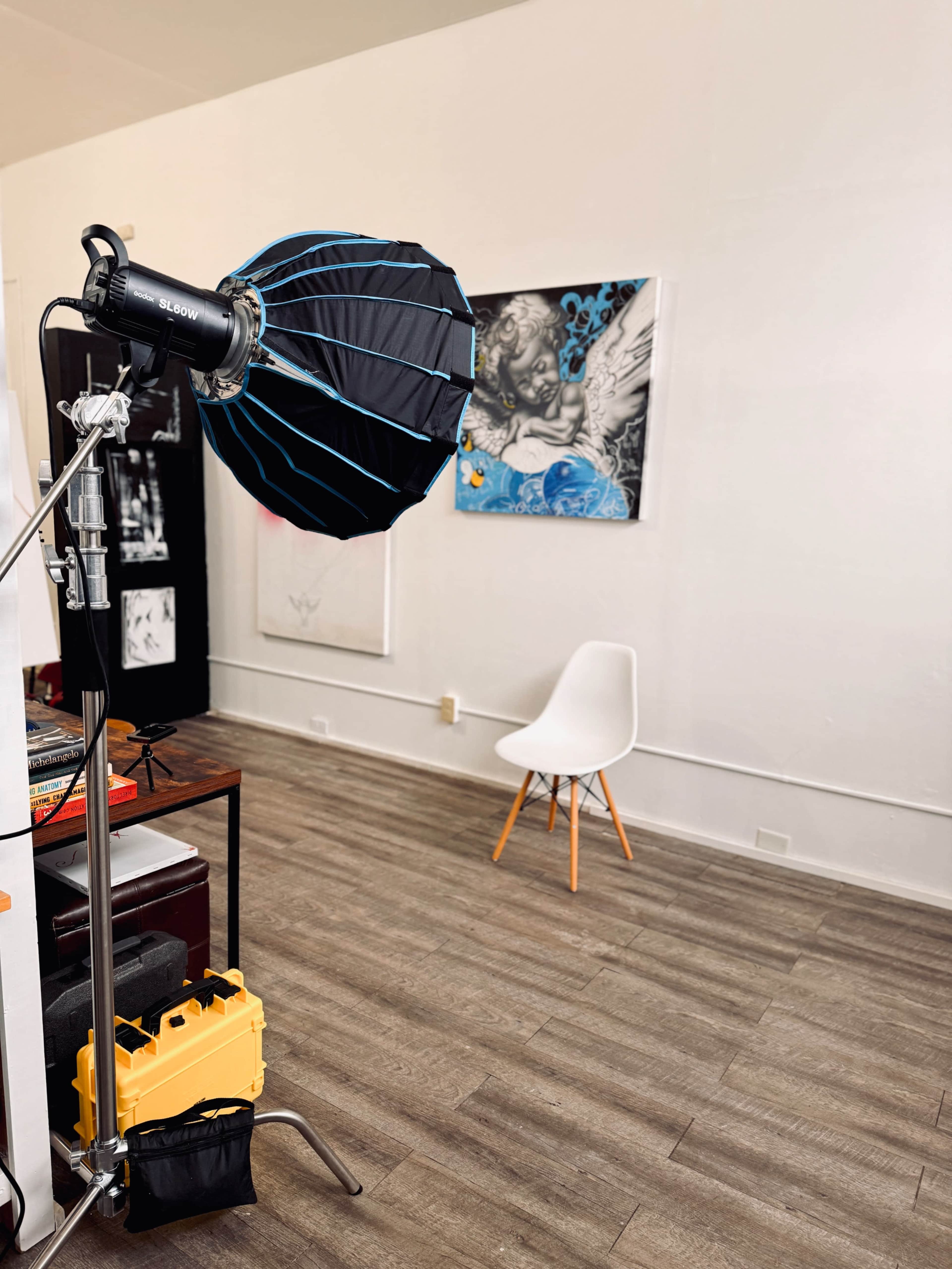 A photography studio setup features a softbox light on a stand beside a wooden table, with a white chair and artwork on the wall in the background.