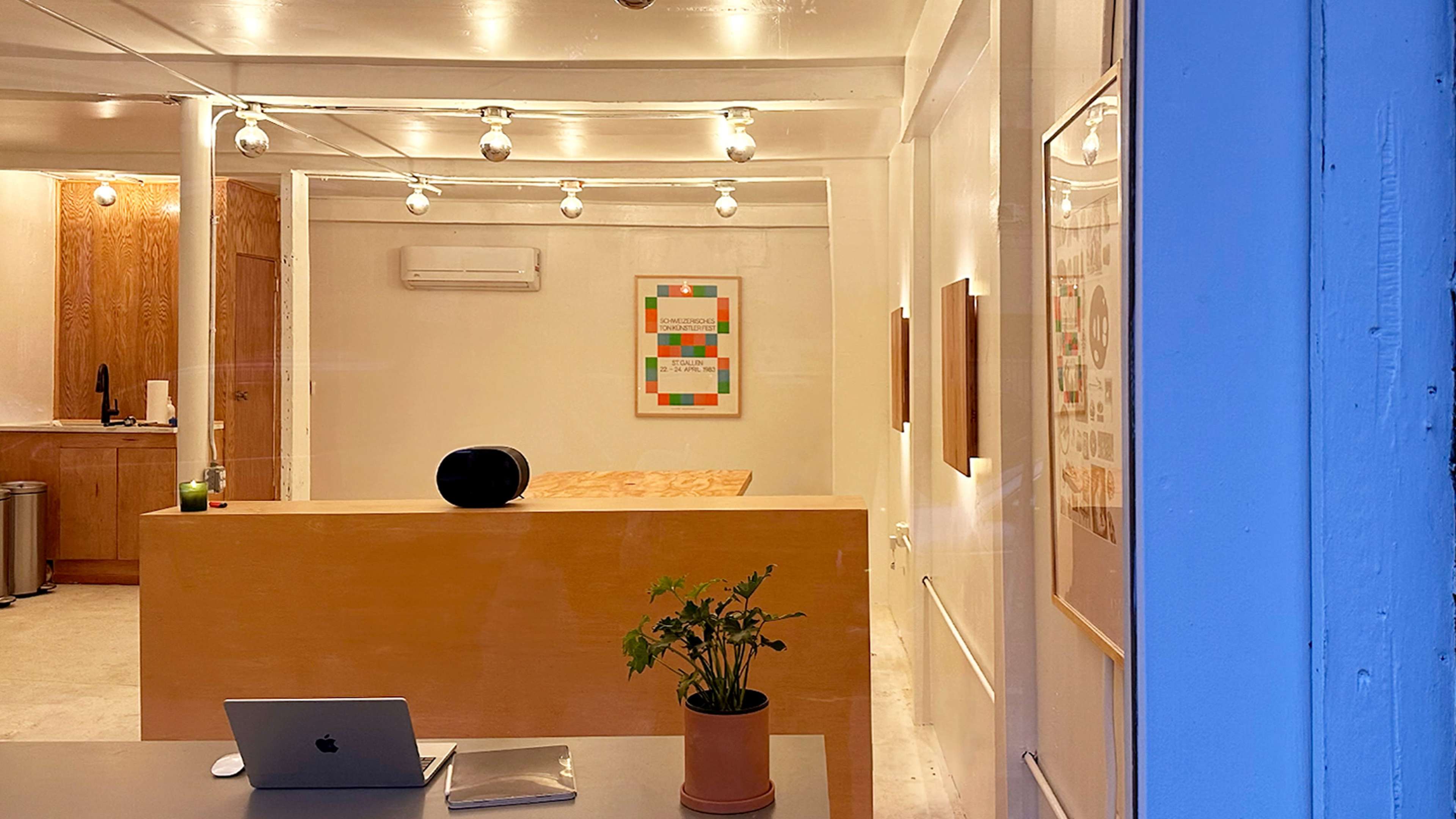 A simple interior features a wooden reception desk, a laptop on a table, and various pieces of artwork on the walls, illuminated by overhead lights.