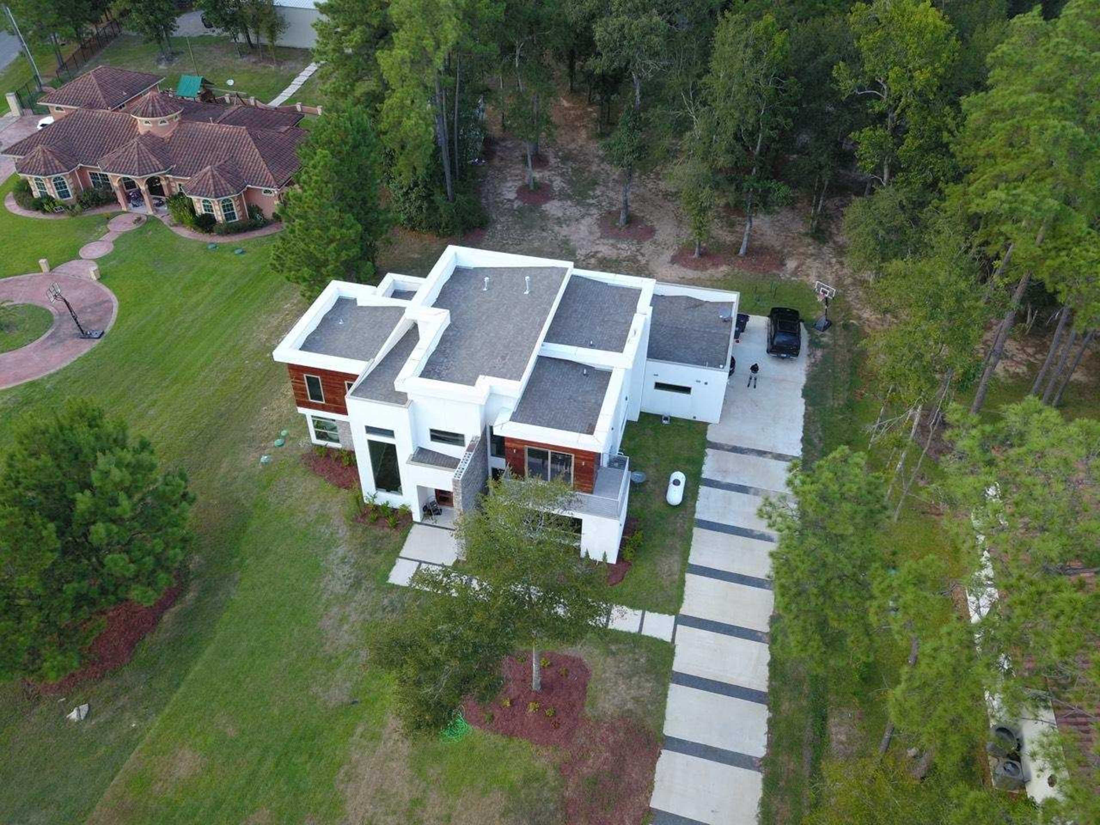 A modern white house located in a wooded area, featuring a driveway with a few parked cars and neighboring homes in the background.