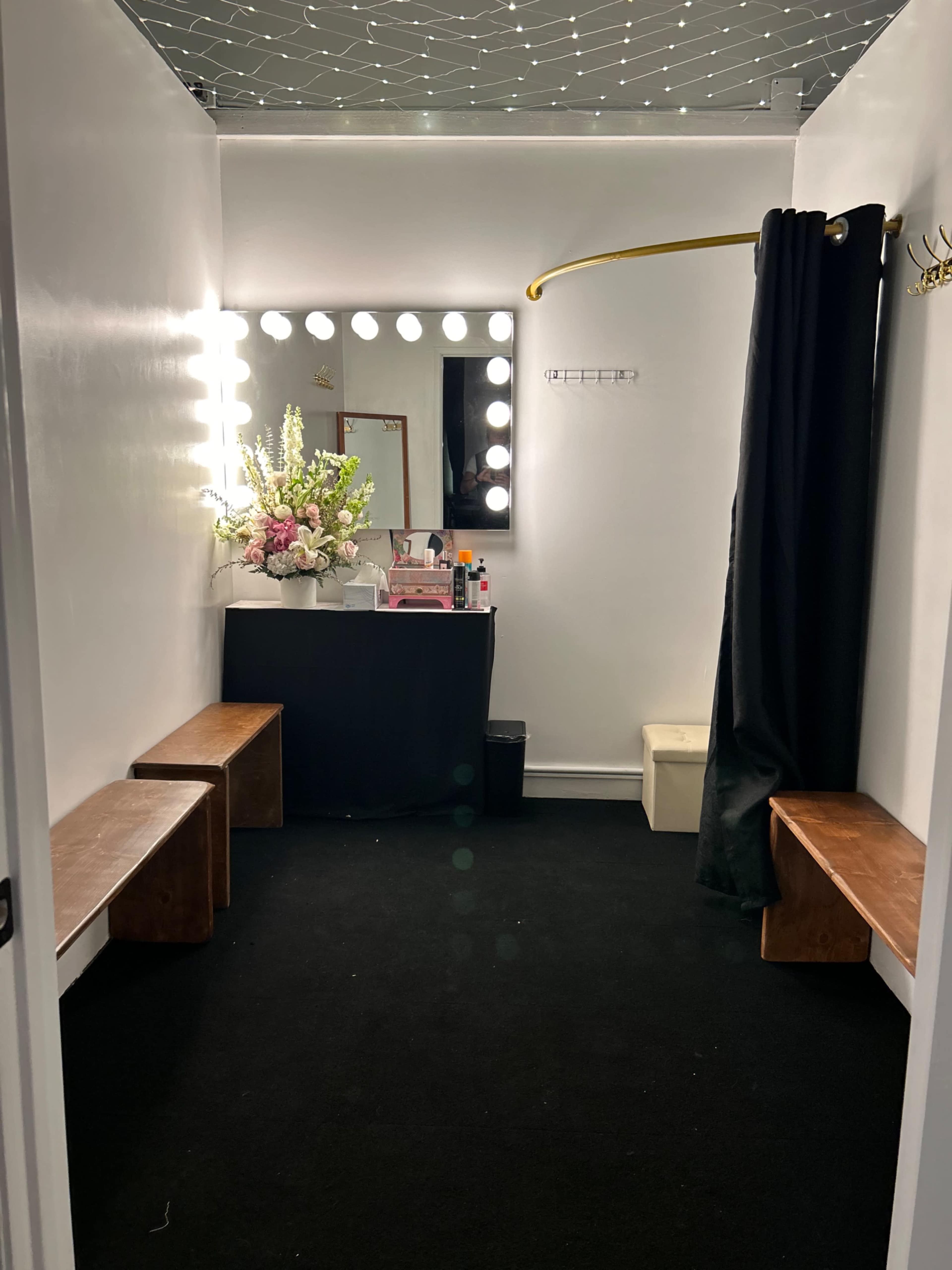 A tidy dressing room with a large mirror framed by bright lights, black benches, and a floral arrangement on a makeup table.