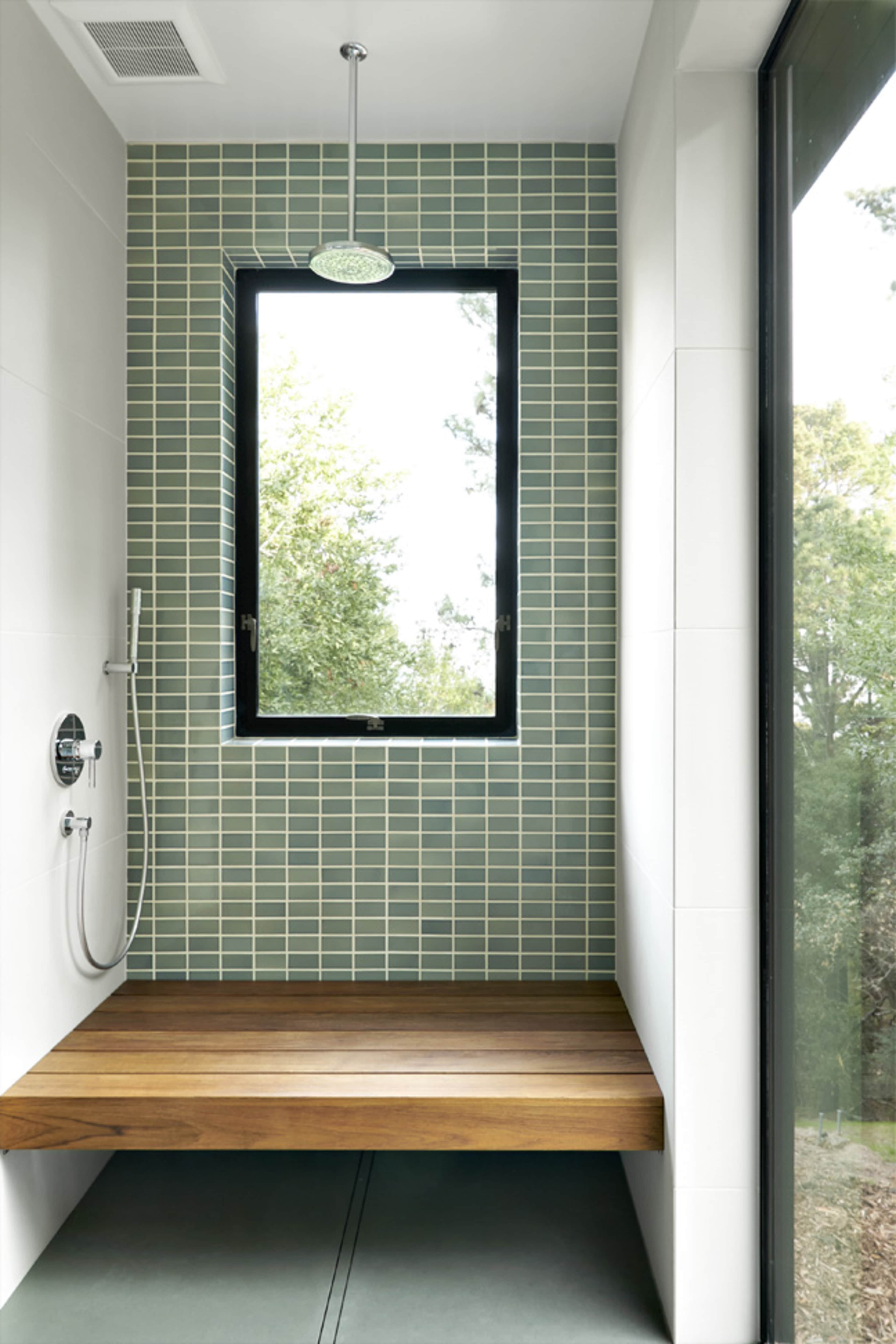 A modern shower space with green tiled walls, a wooden bench, and a large window overlooking greenery.