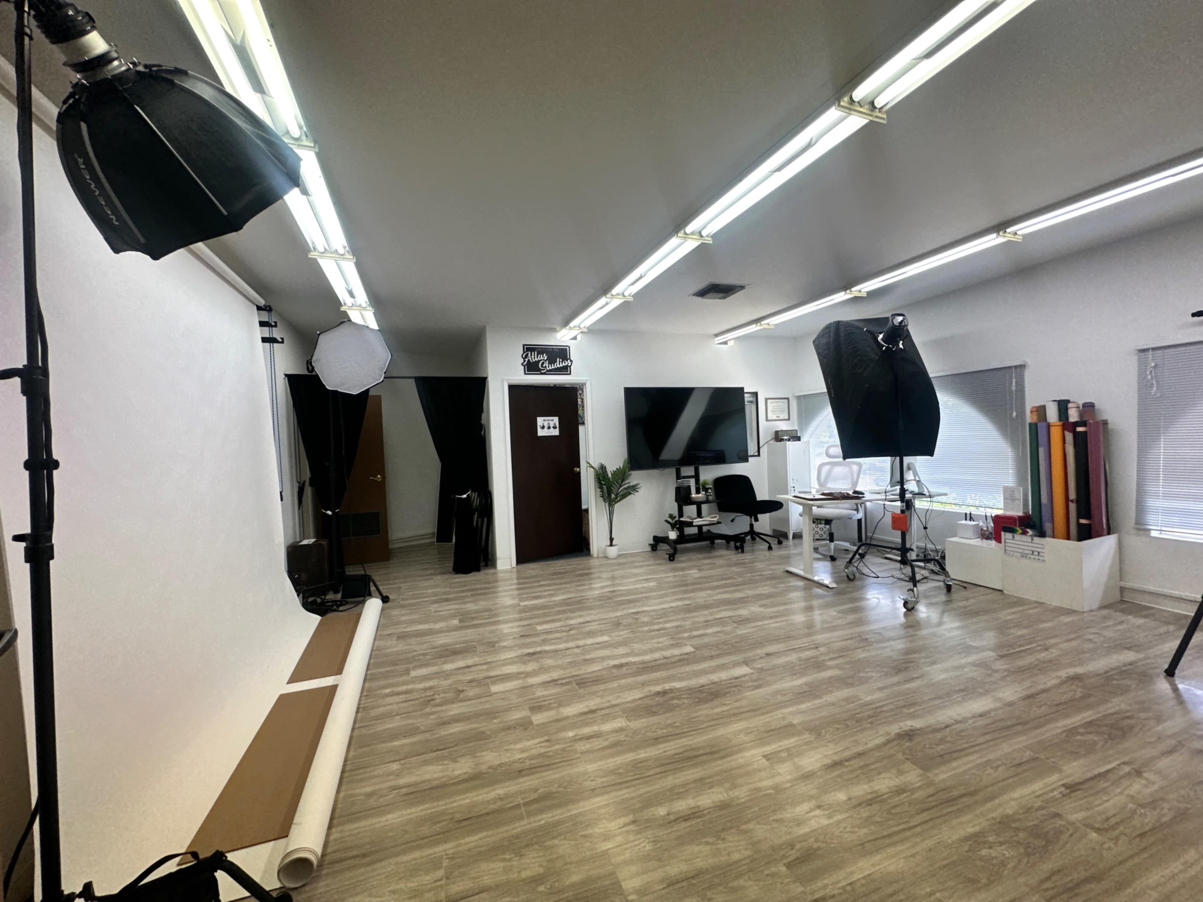 A photography studio with lights, a backdrop, a large television, and a studio chair arranged in a spacious area with wooden flooring.