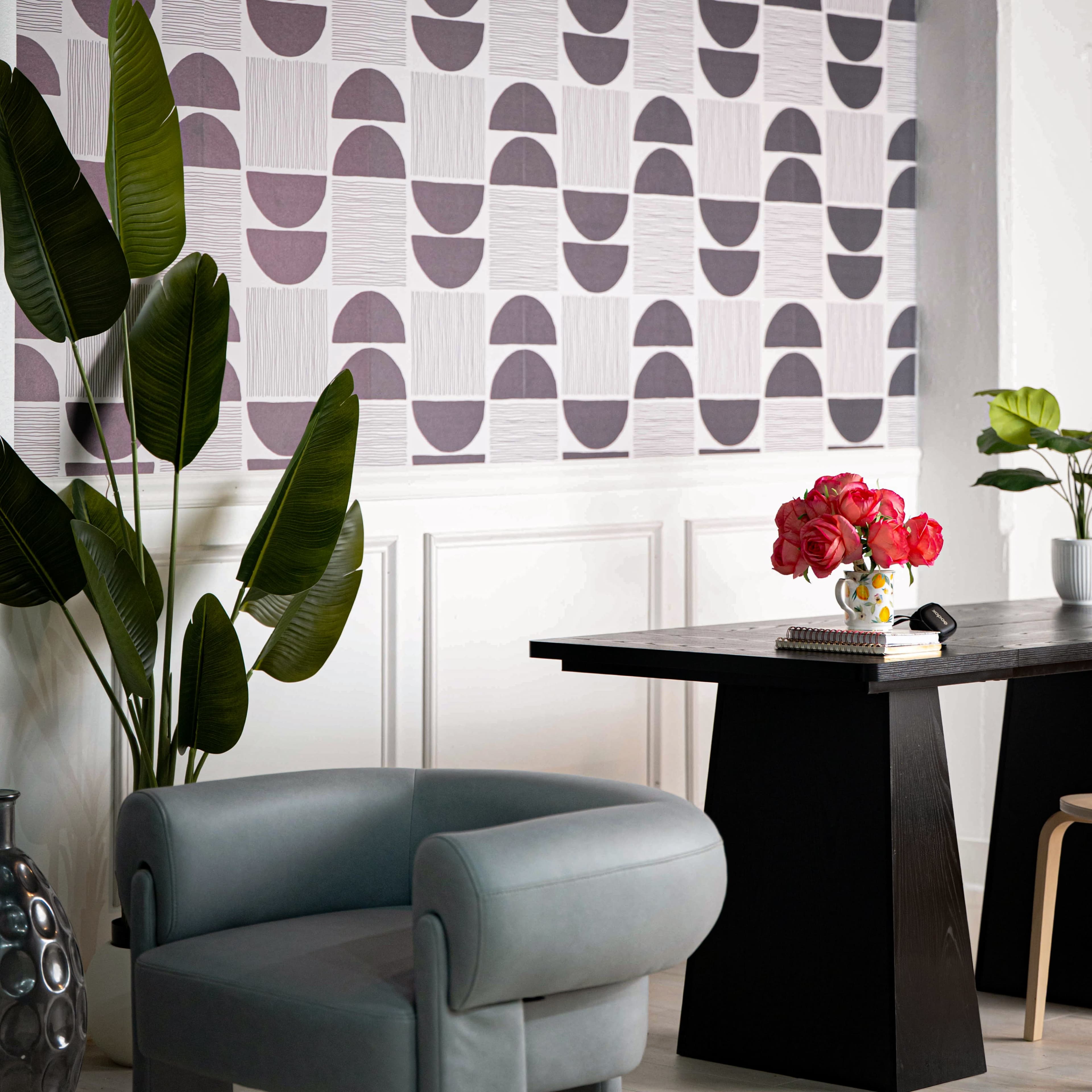 The image features a modern interior with a patterned wallpaper, a dining table, a chair, and decorative plants and flowers.