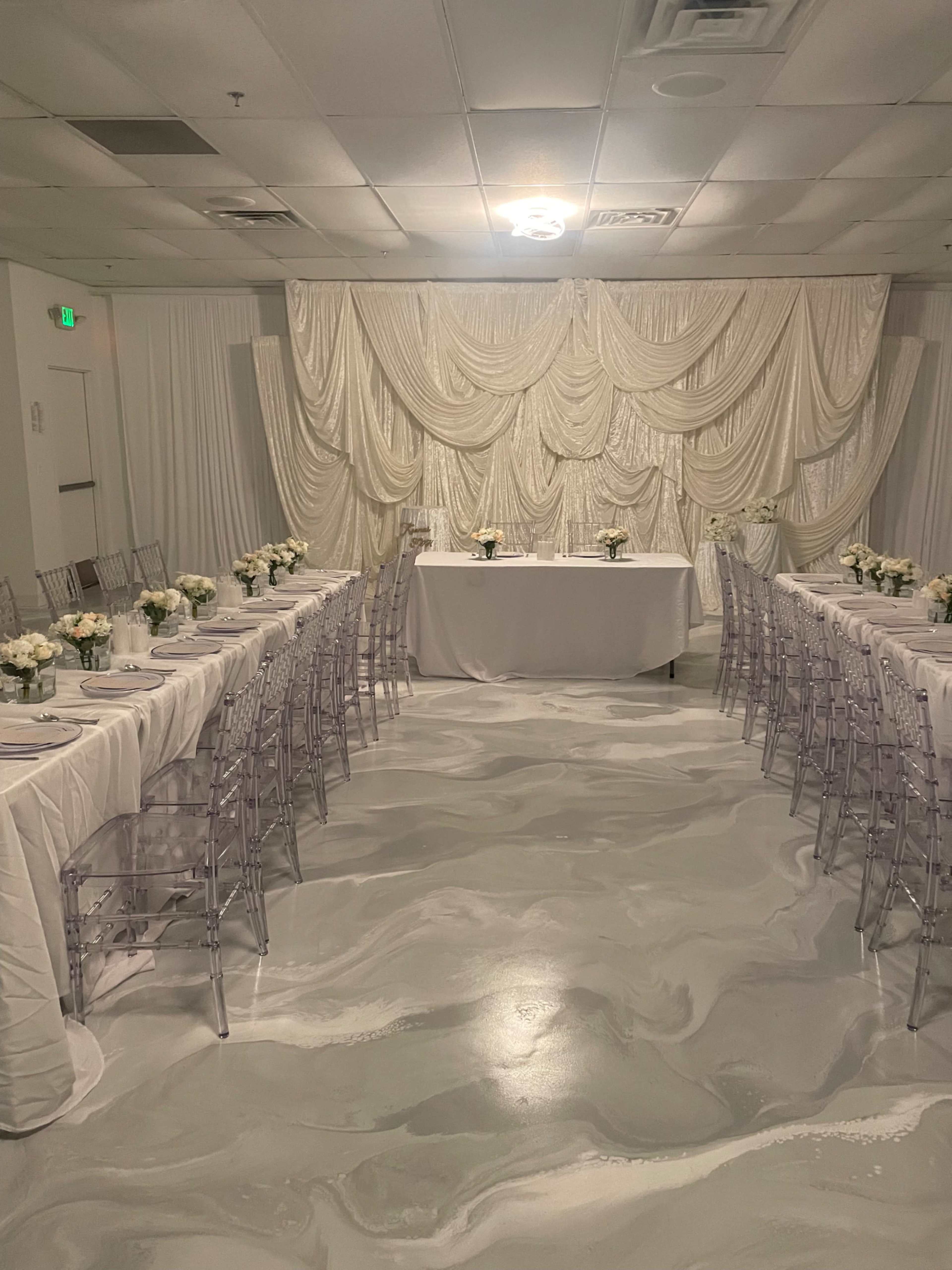 The image shows a banquet room set up with two long tables adorned with white tablecloths and floral centerpieces, surrounded by transparent ghost chairs, with a textured white backdrop draping behind the head table.