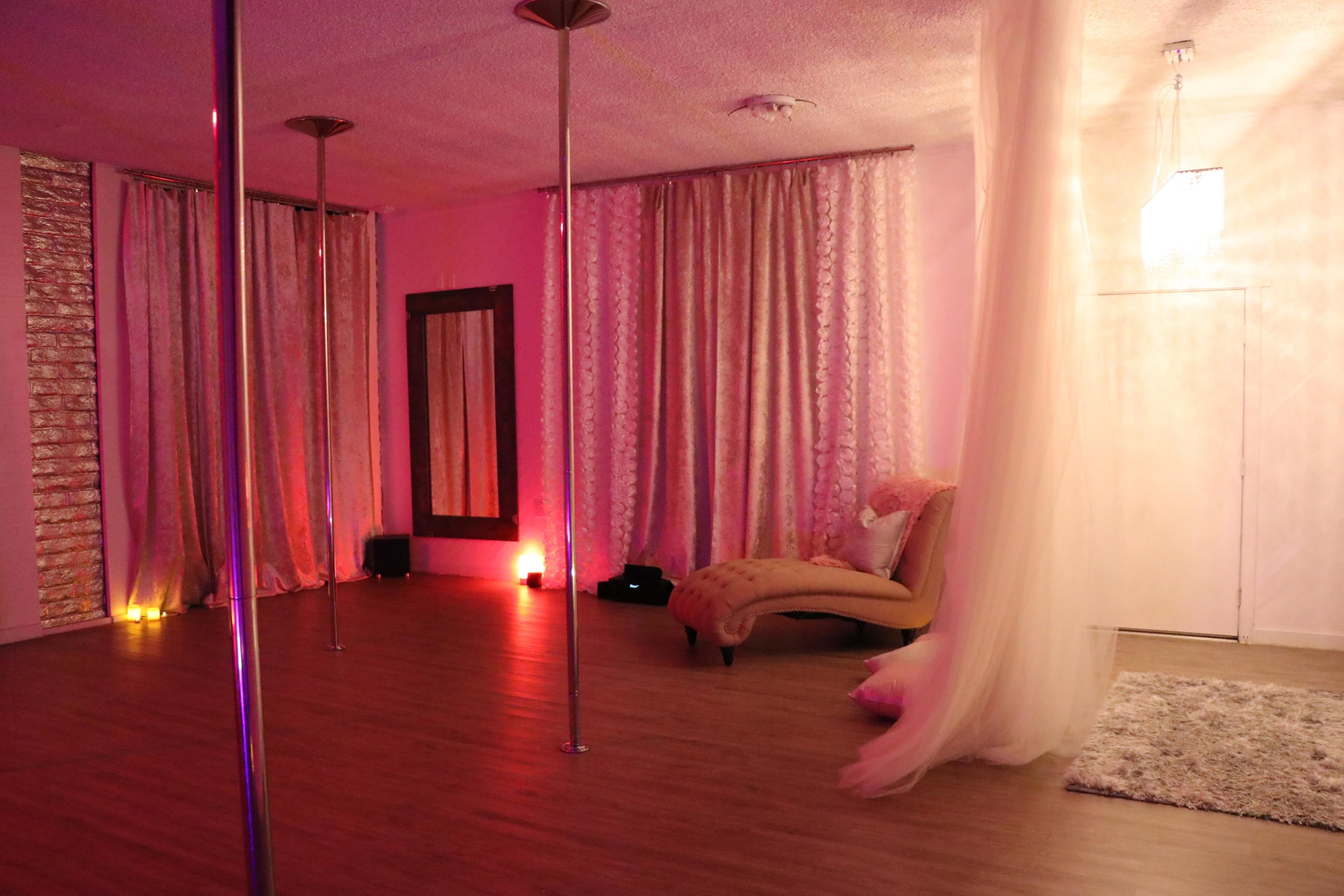 A dance studio with two poles, soft lighting, draped curtains, and a chaise lounge.