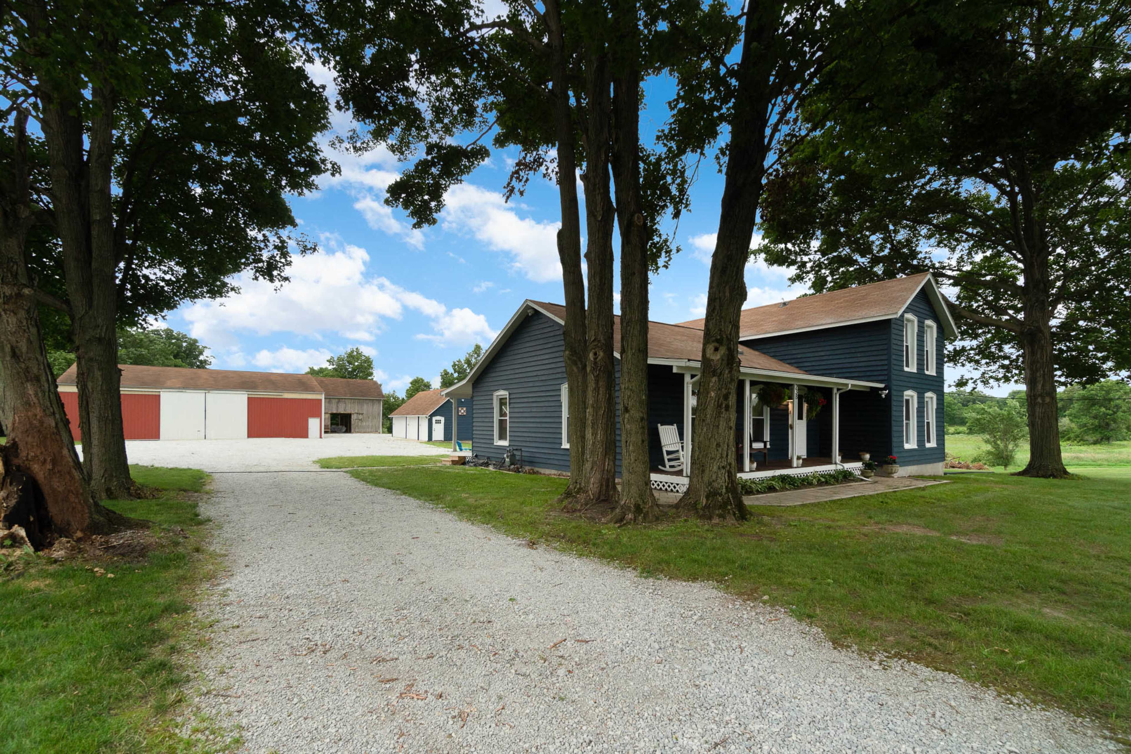 A blue two-story house is set beside a gravel driveway leading to a large red and beige barn, surrounded by green grass and trees.