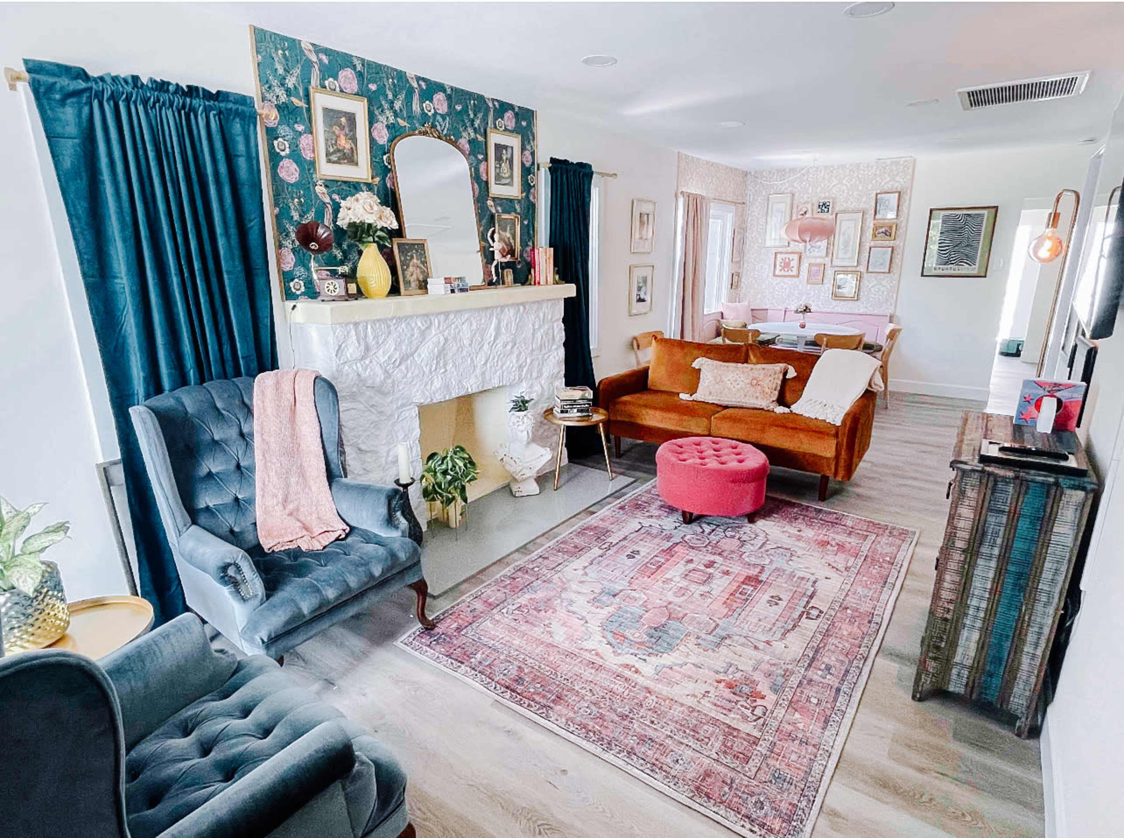 The room features a living area with a stone fireplace, two upholstered chairs, a brown sofa, a pink ottoman, and a decorative rug.