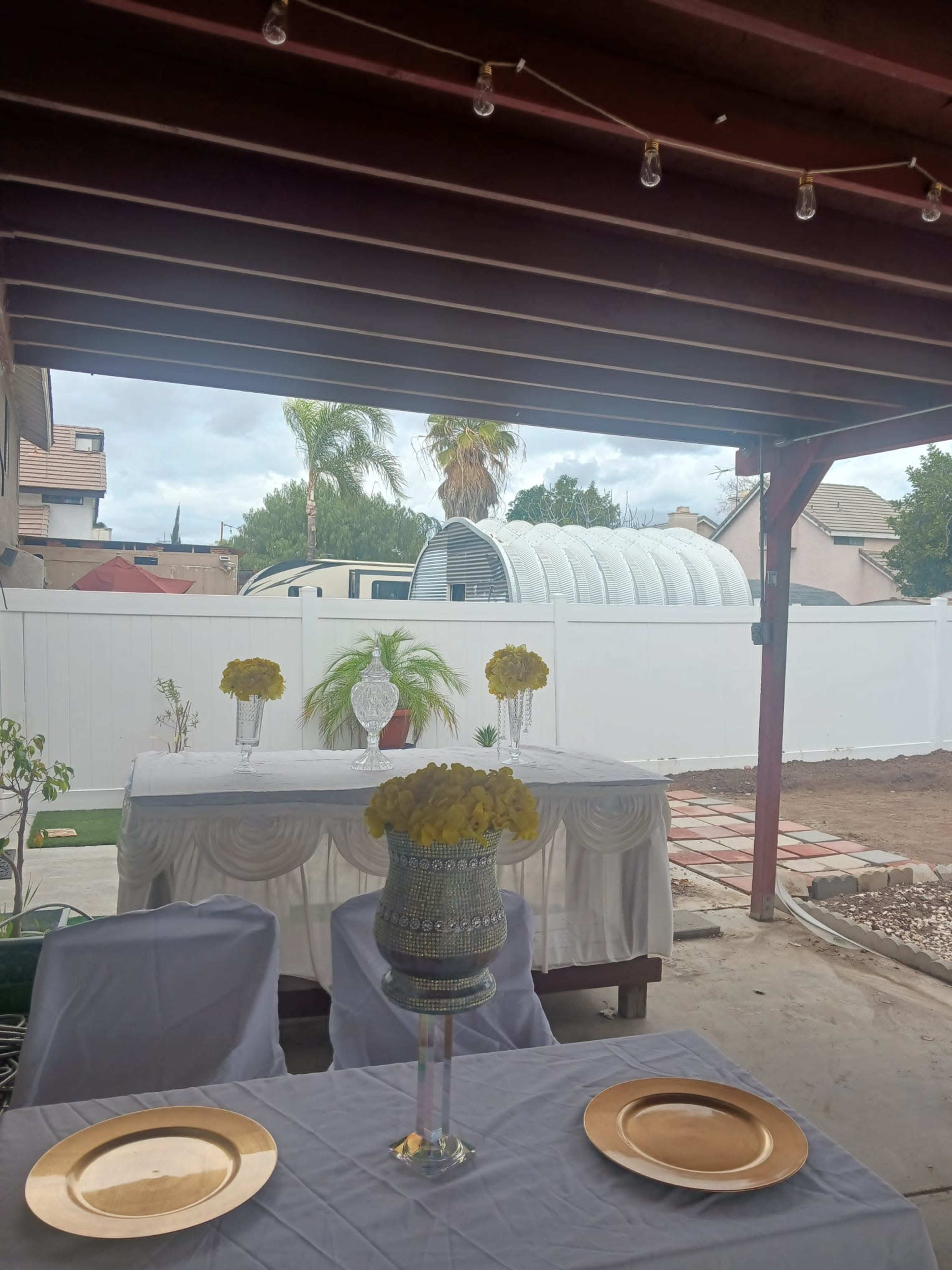 The image shows a covered outdoor area with a dining table set for a meal, featuring decorative flowers, and a white fence in the background.