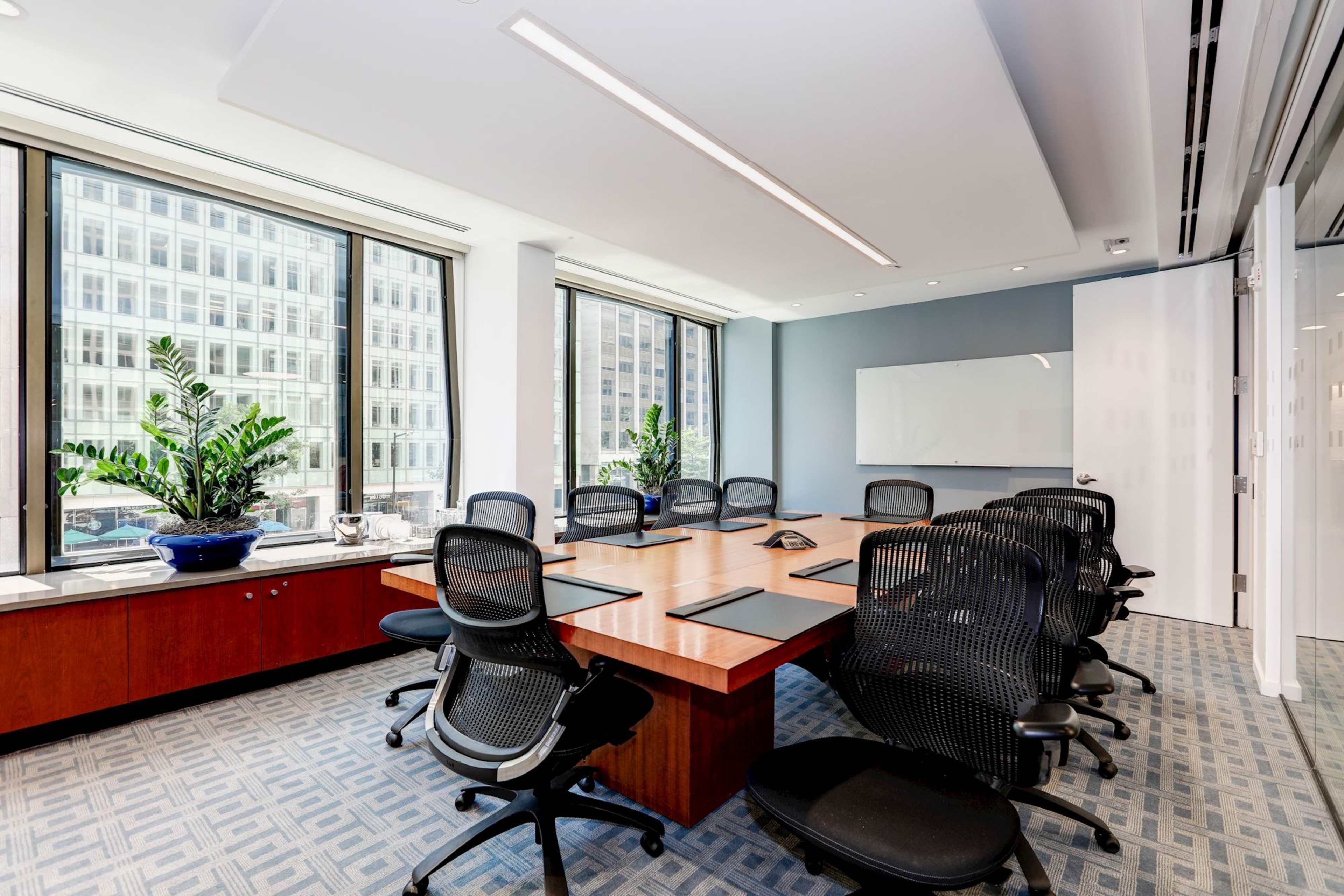 A conference room with a large wooden table surrounded by black ergonomic chairs, featuring large windows that provide a view of a cityscape and potted plants in the corners.