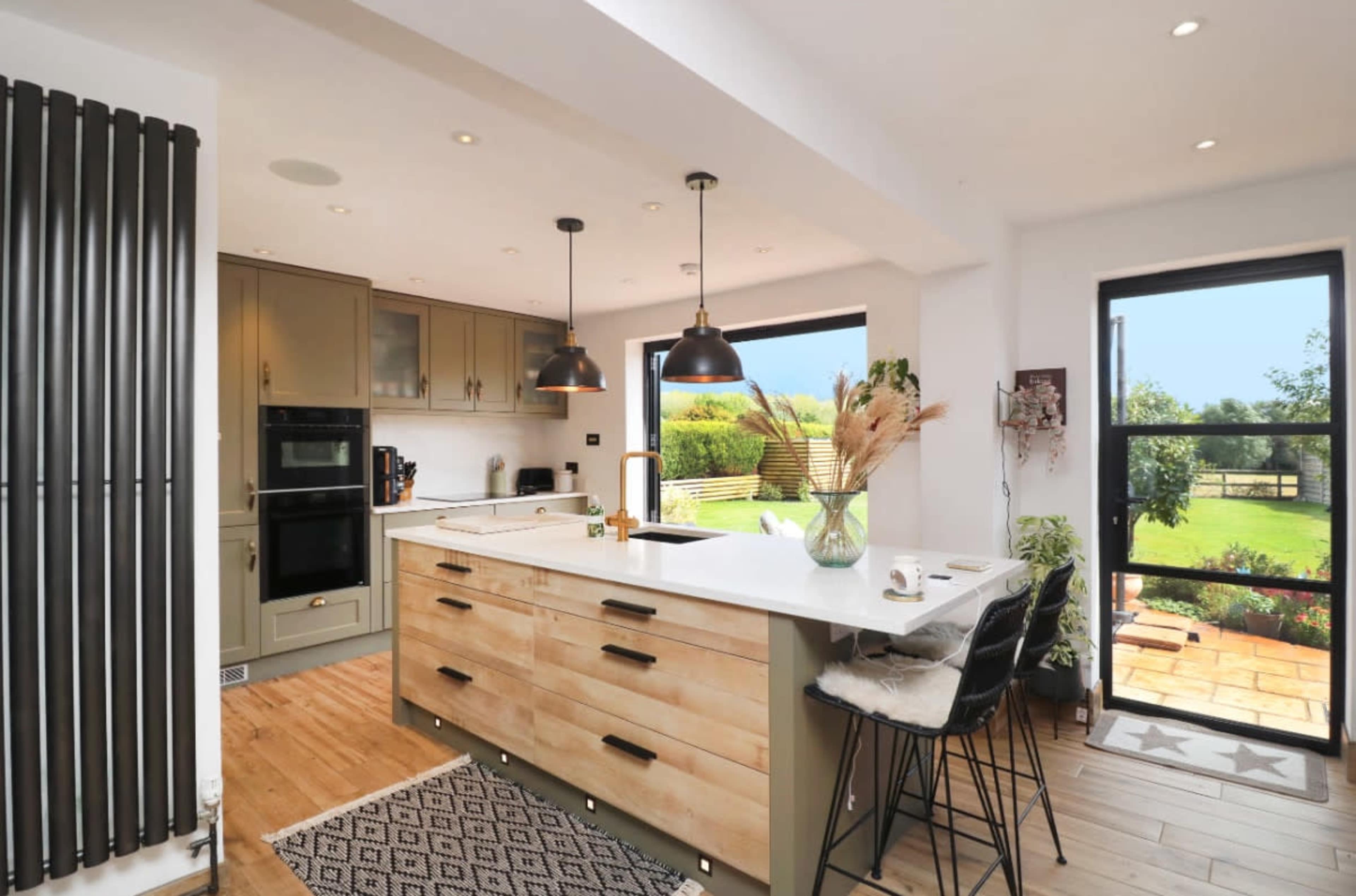 The image shows a modern kitchen with an island, featuring olive green cabinetry, a white countertop, and large windows overlooking a garden.