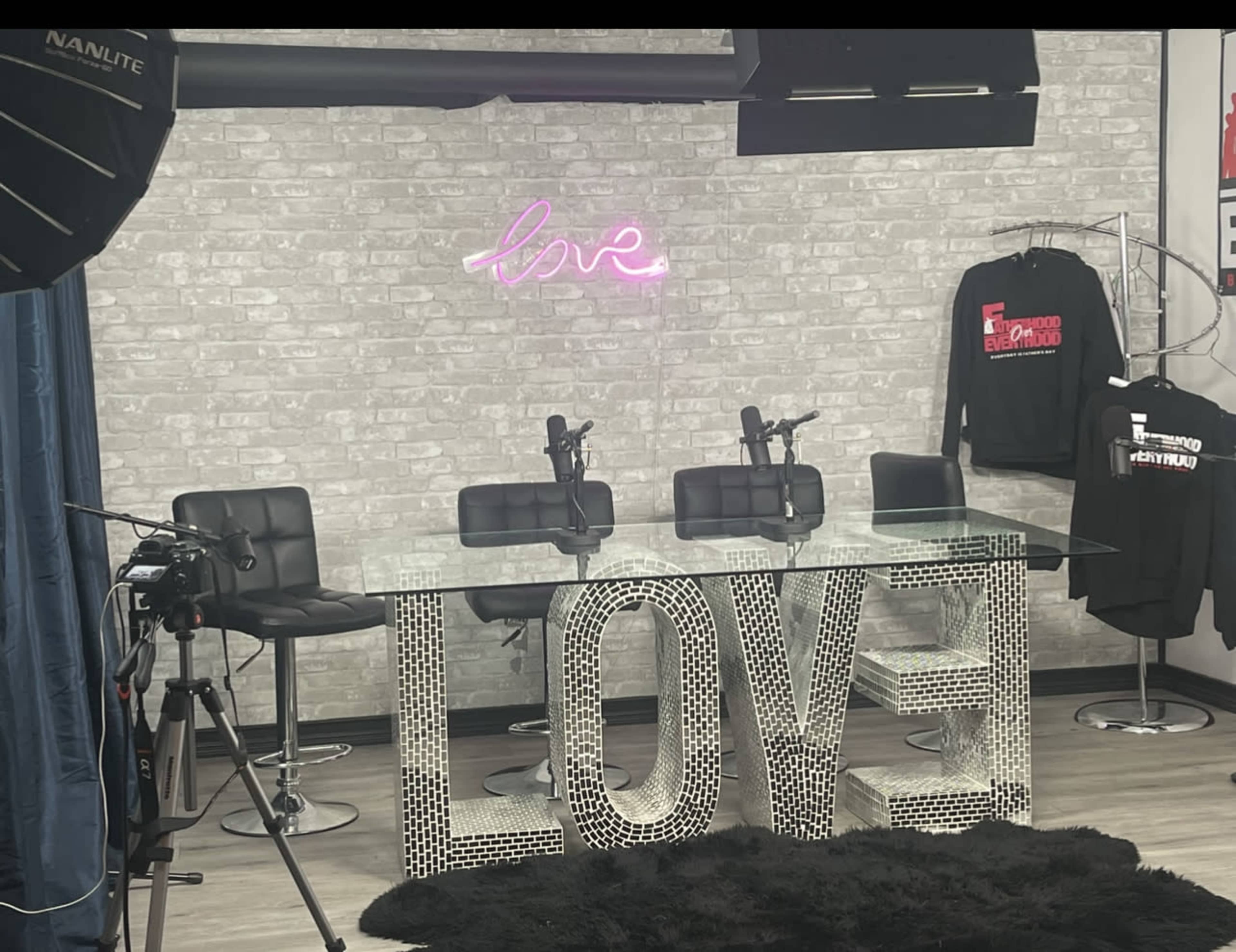 The image shows a studio setup with a glass table featuring the word "LOVE" made of reflective materials, surrounded by black chairs and a neon sign on the wall.