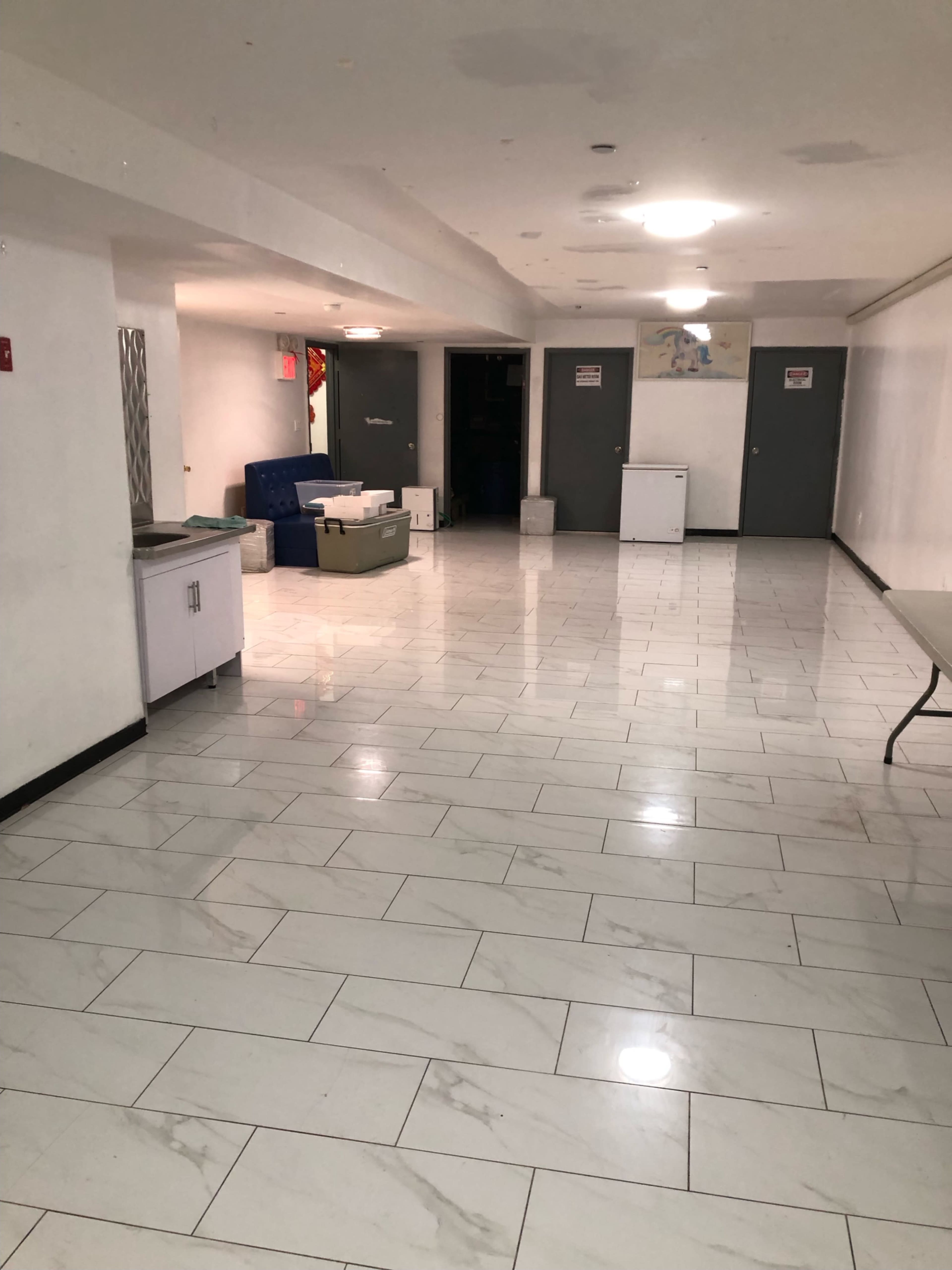 The image shows a spacious, empty room with tiled flooring, featuring a table and some furniture, along with several closed doors.