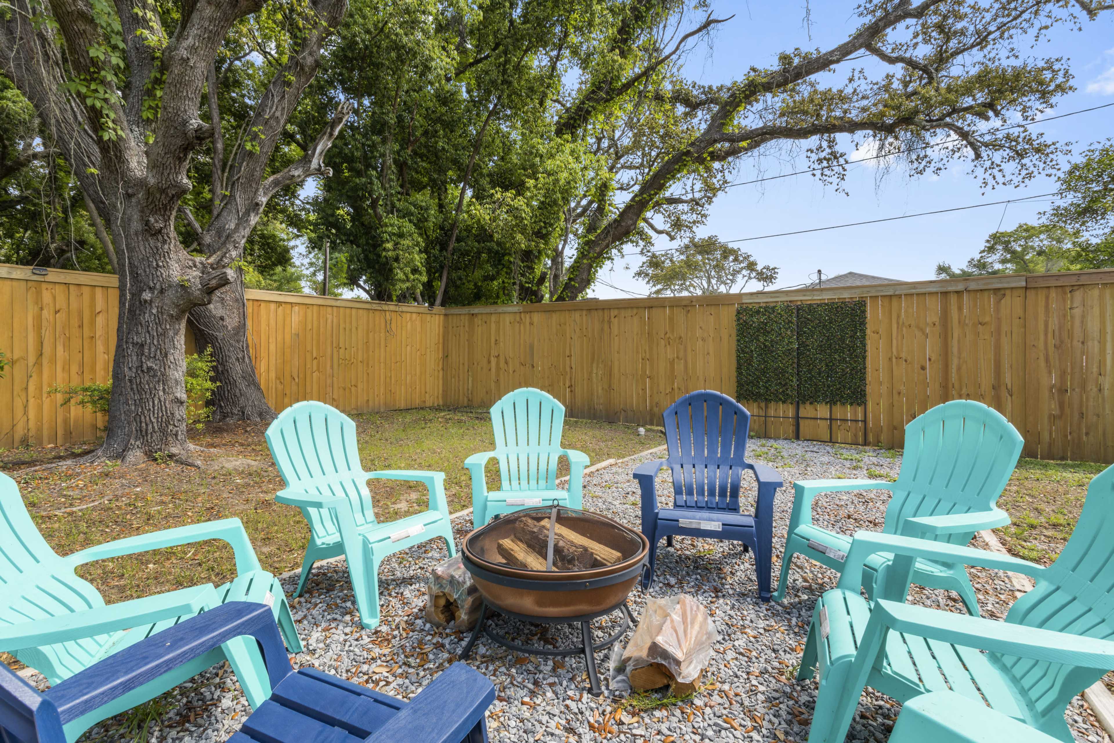 A fire pit surrounded by eight colorful adirondack chairs is set in a grass area enclosed by a wooden fence.
