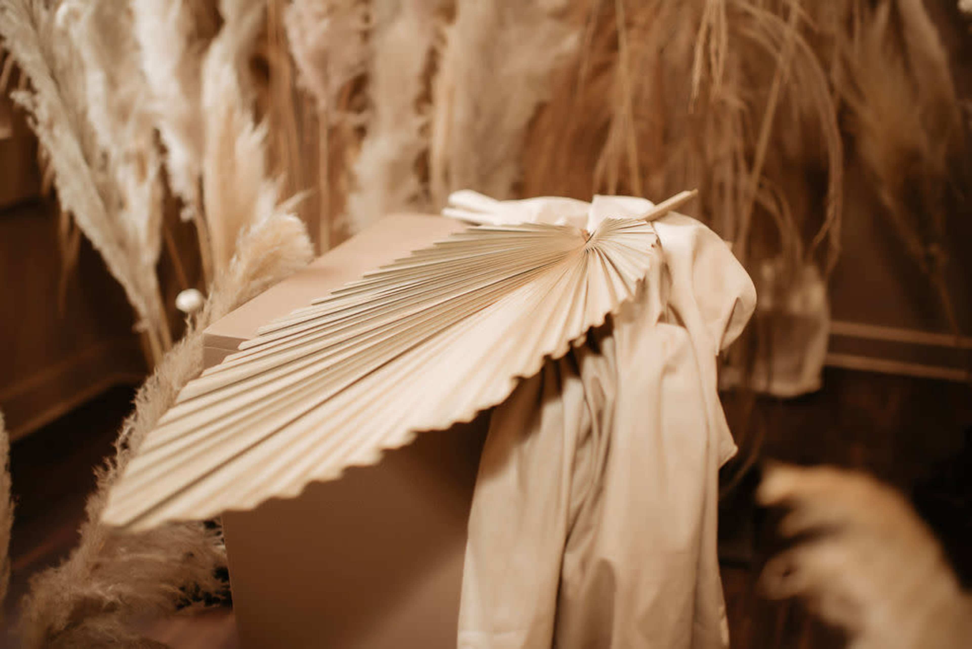 A large, decorative fan rests on a draped cloth on a table surrounded by dried pampas grass.