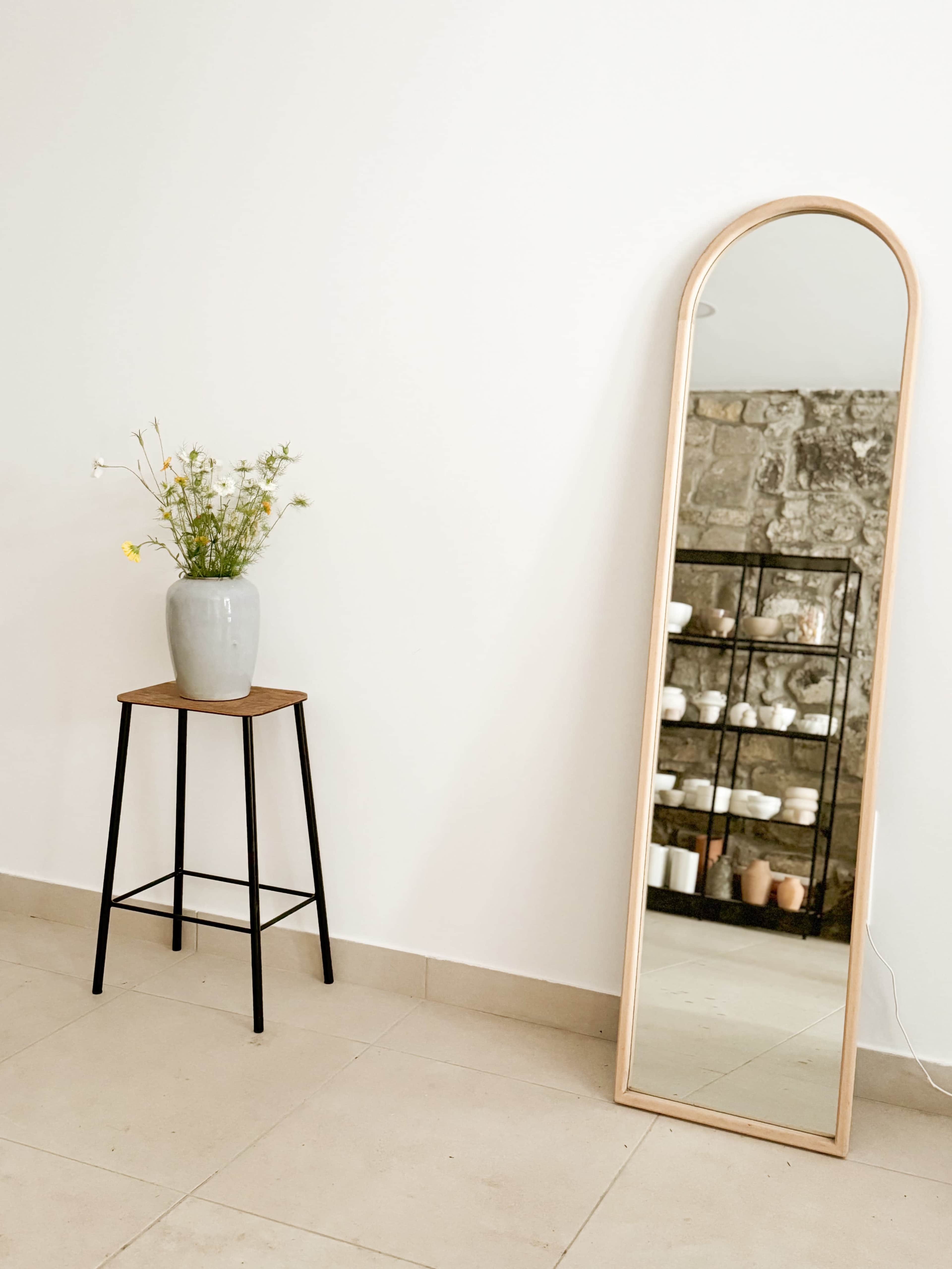A tall, arched mirror stands against a white wall beside a potted flower arrangement on a black metal stool, with a stone wall shelf displaying various objects in the background.