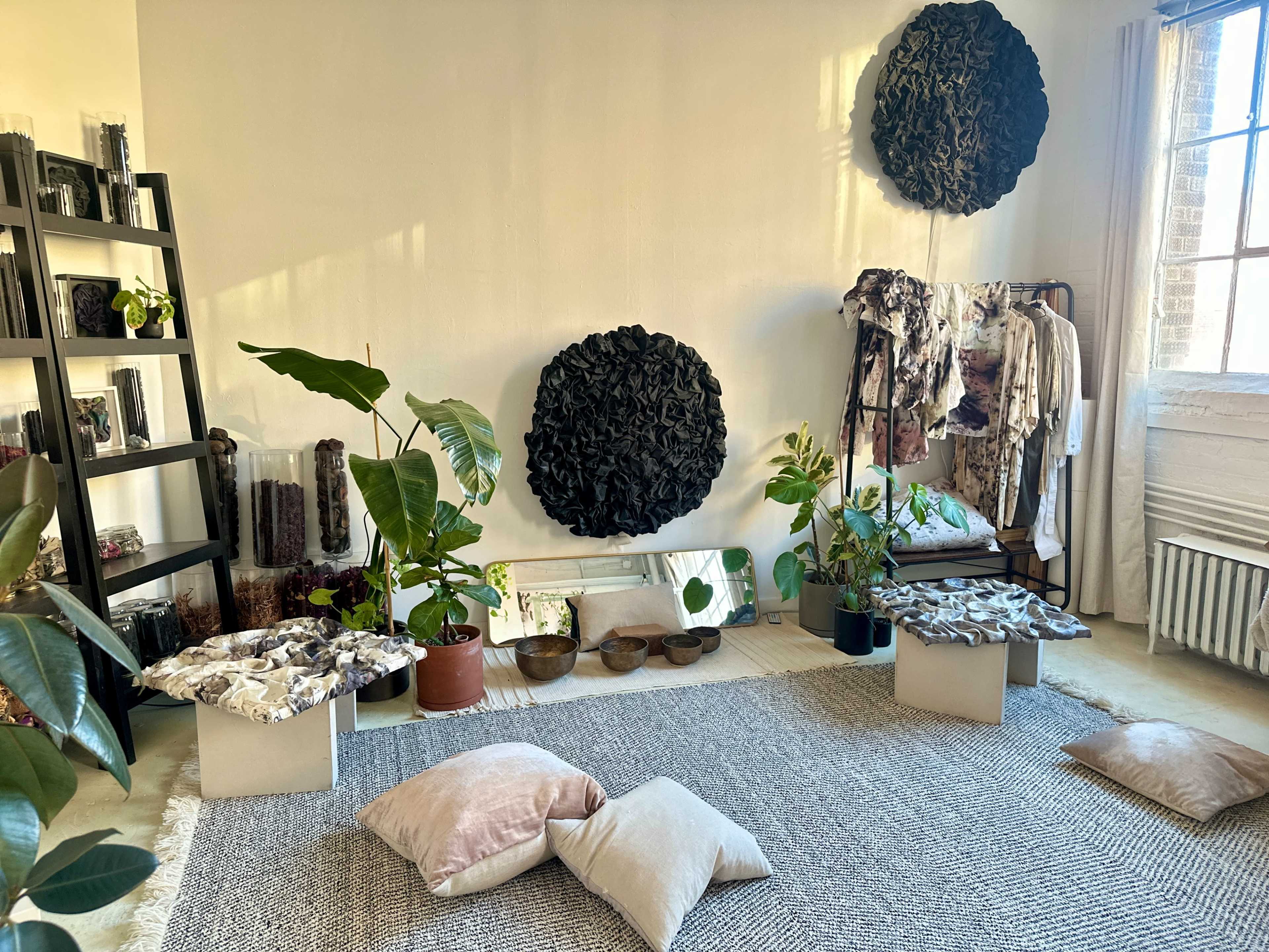 The image depicts a stylish interior space featuring decorative black wall art, potted plants, and seating cushions arranged on a textured rug.