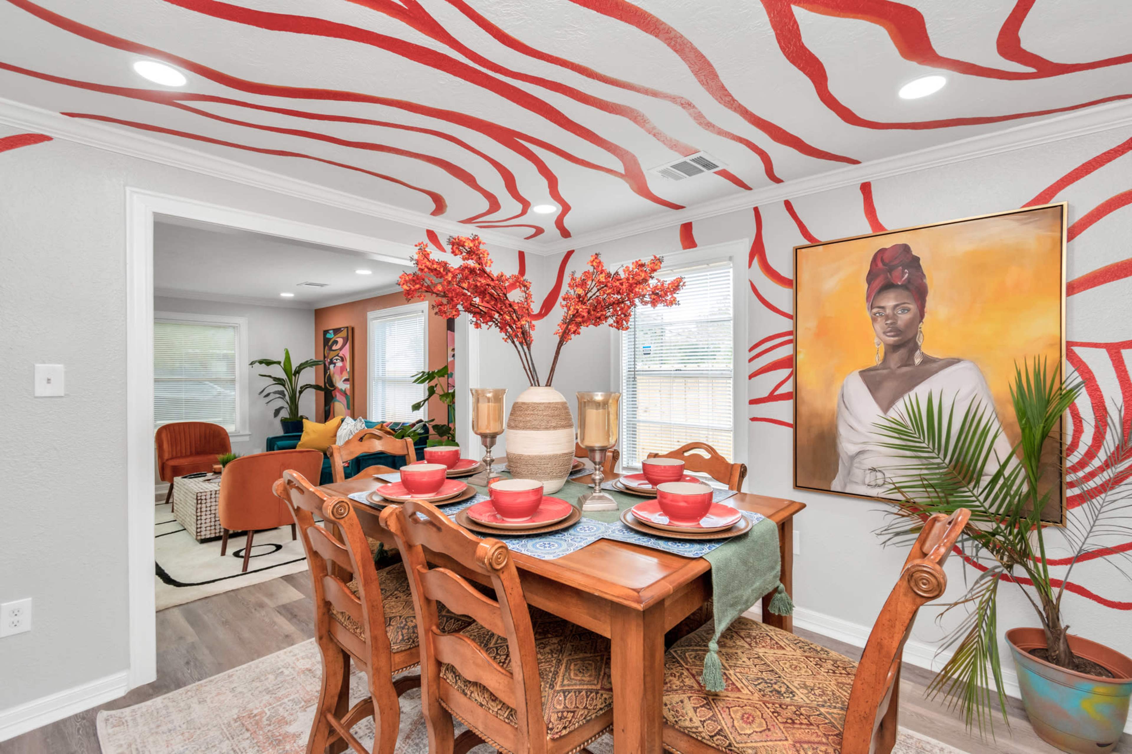 A dining area features a wooden table set with red dishes, surrounded by vibrant wall art and decorative plants.