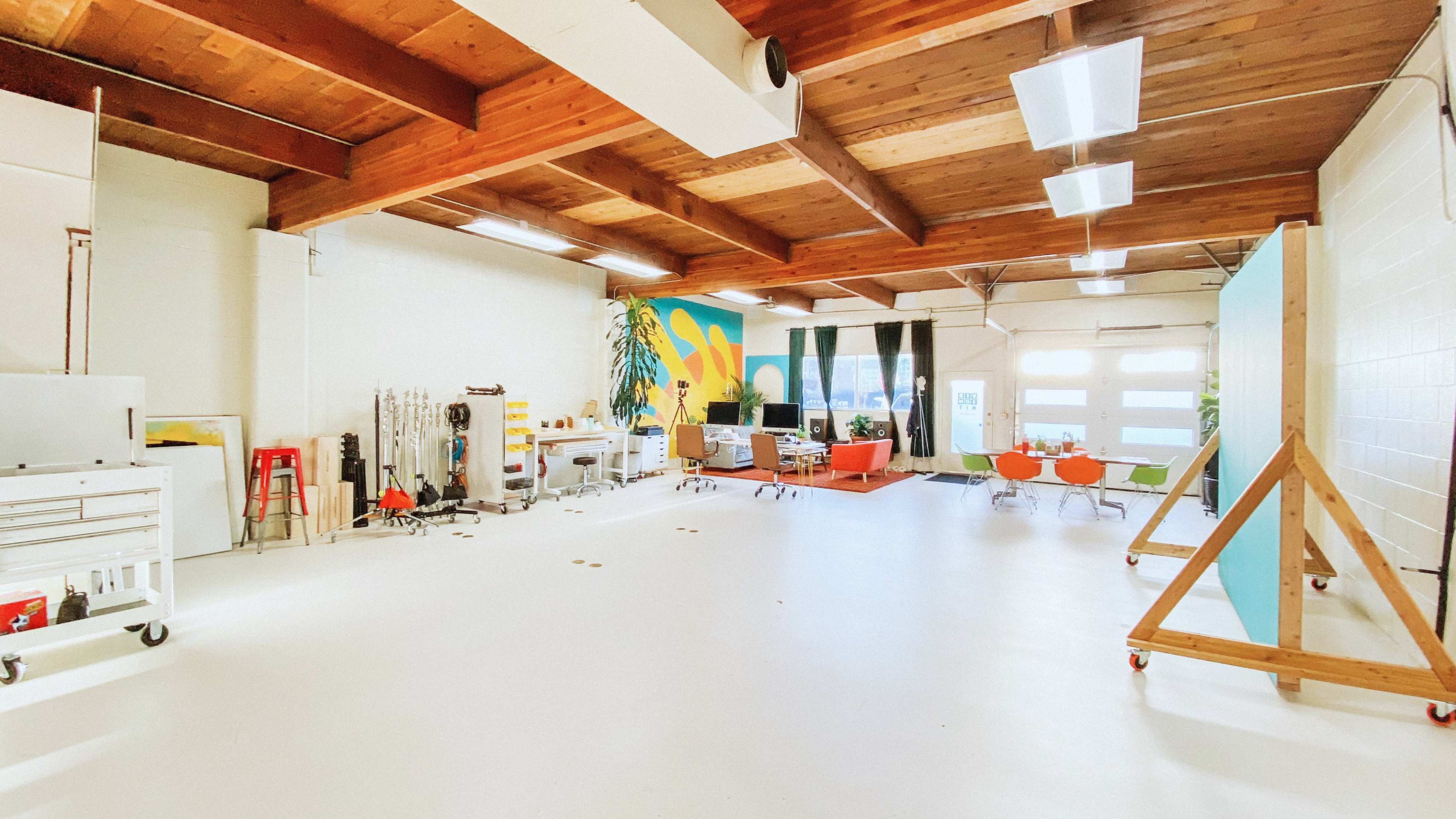 A spacious, well-lit studio with wooden beams, colorful walls, and various furniture arrangements, including seating areas and workspaces.