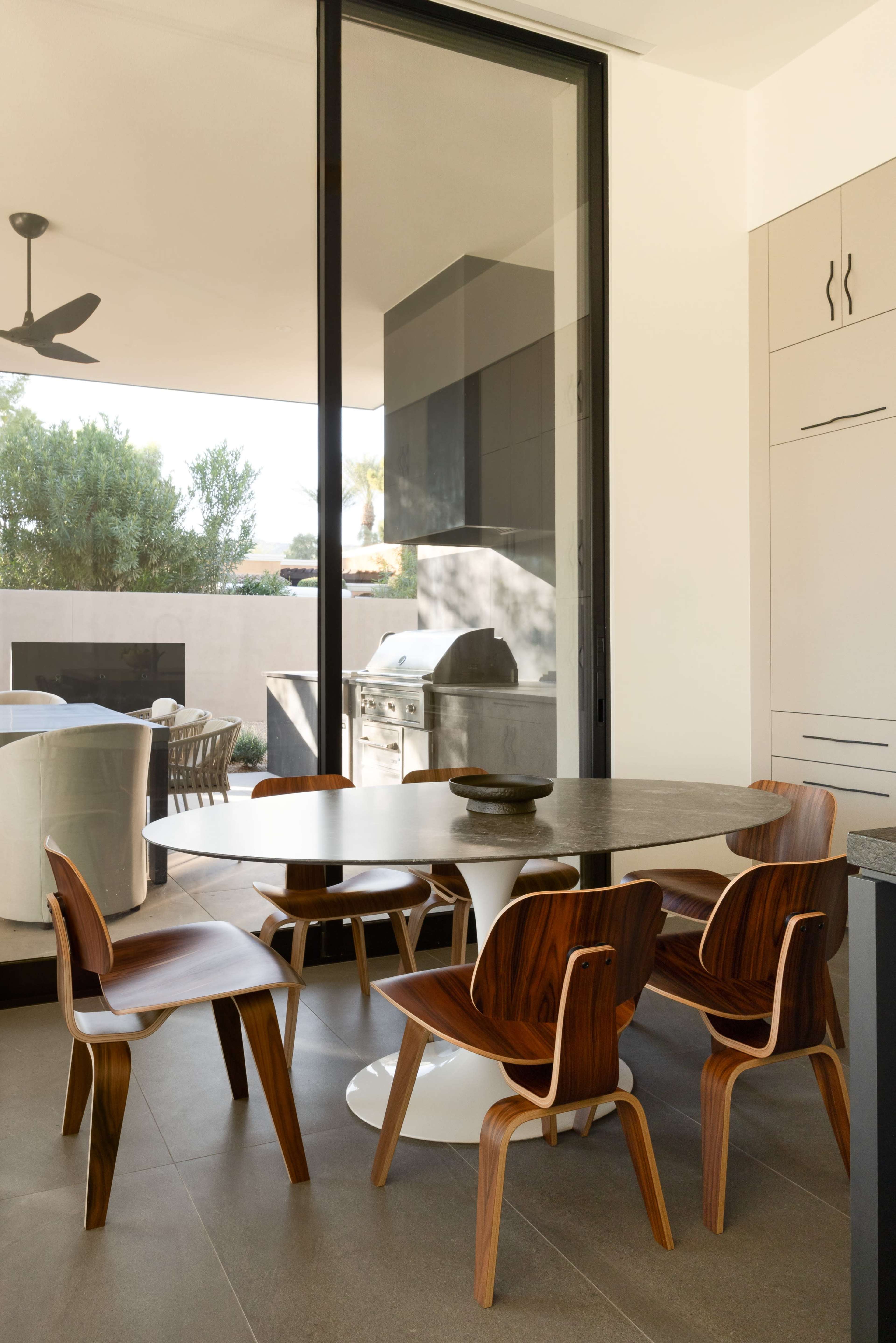 A modern kitchen features a round table with wooden chairs, surrounded by large windows that provide a view of an outdoor cooking area.