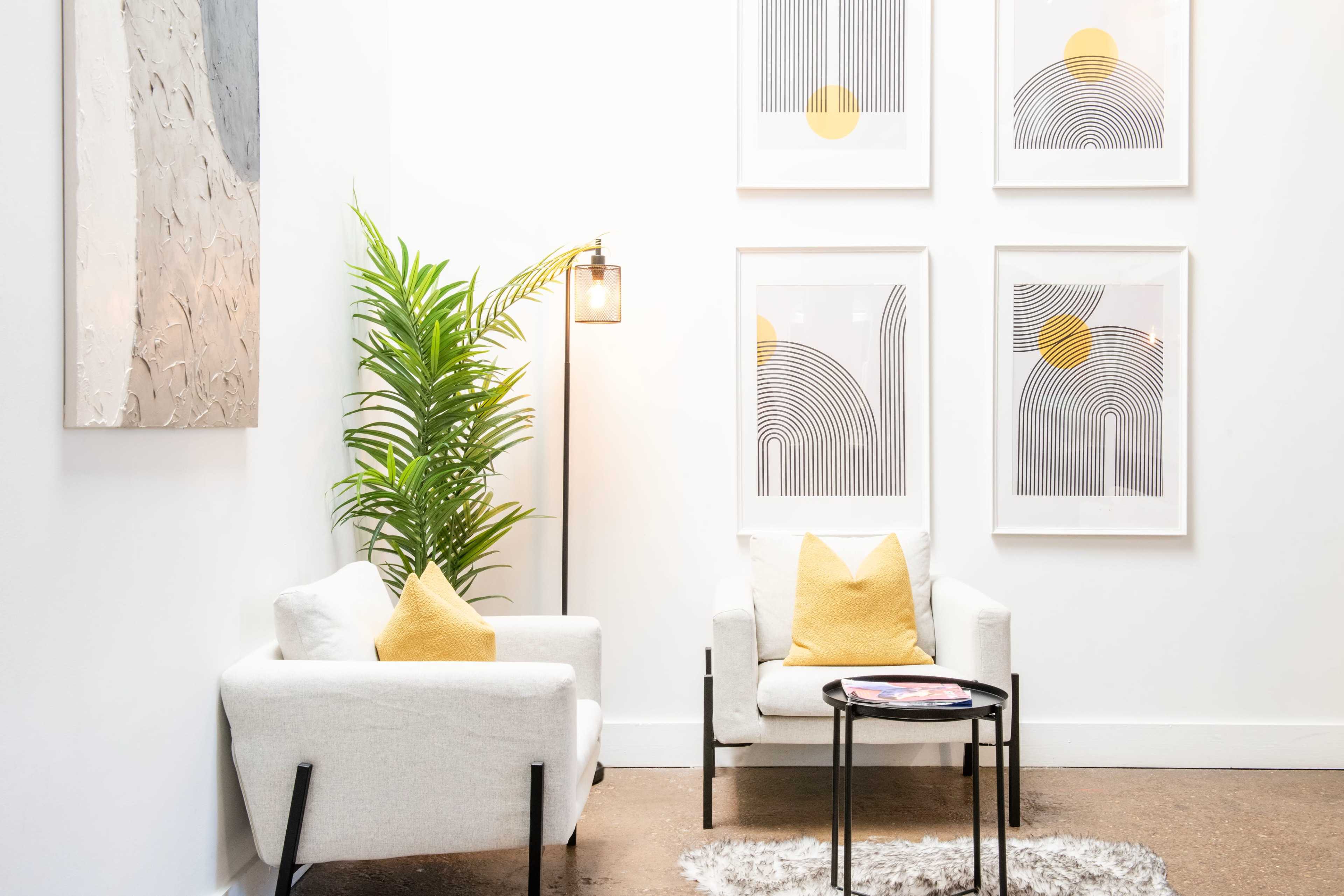 A bright, minimalist interior featuring two white armchairs with yellow cushions, a floor lamp, a potted plant, and three framed abstract artworks on the wall.