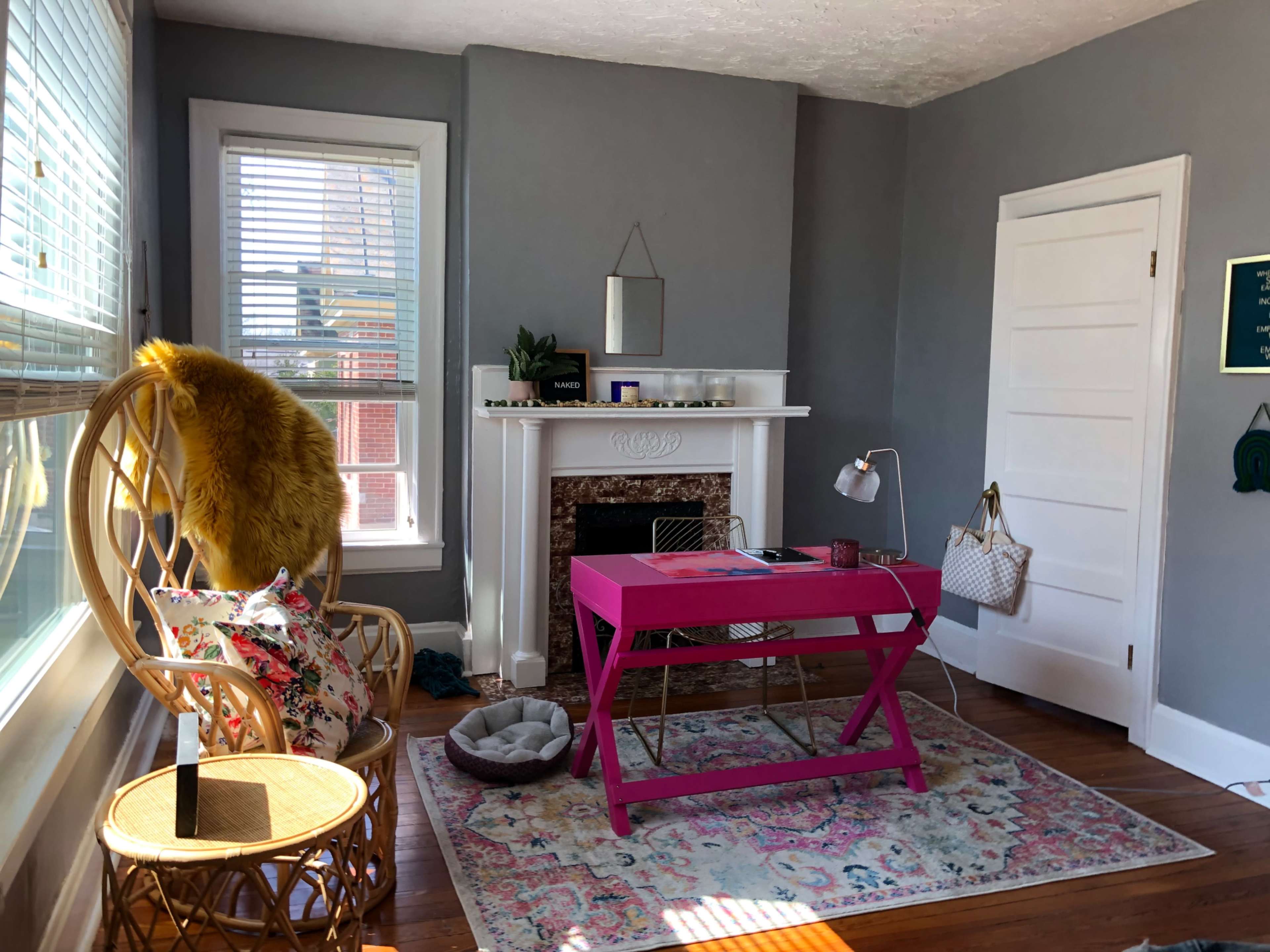 The image shows a brightly lit room with a pink desk, a rattan chair, and a decorative rug in front of a fireplace.