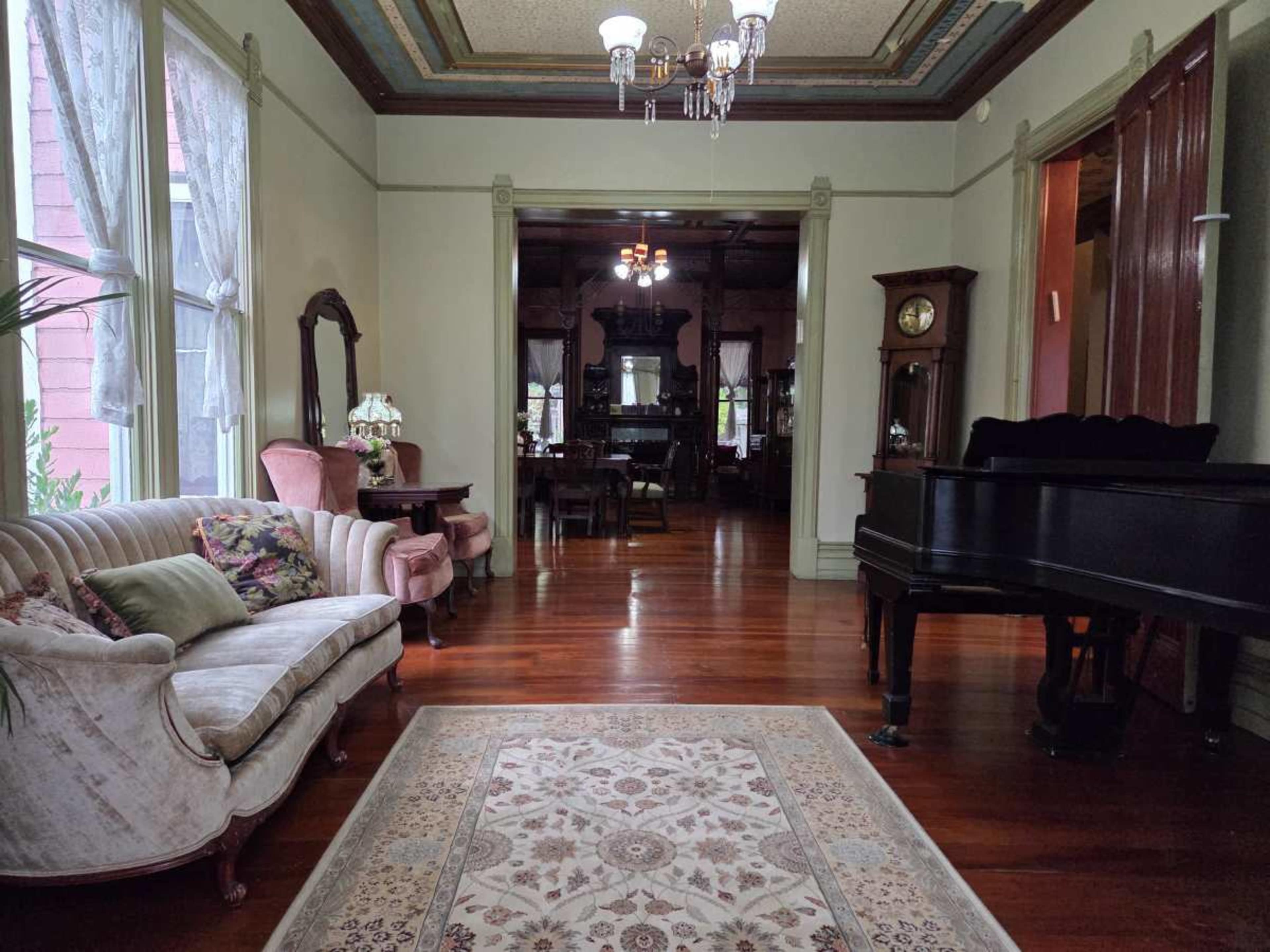 The image shows a spacious, elegantly decorated room with a plush sofa, a grand piano, and a view into a dining area adorned with vintage furniture.