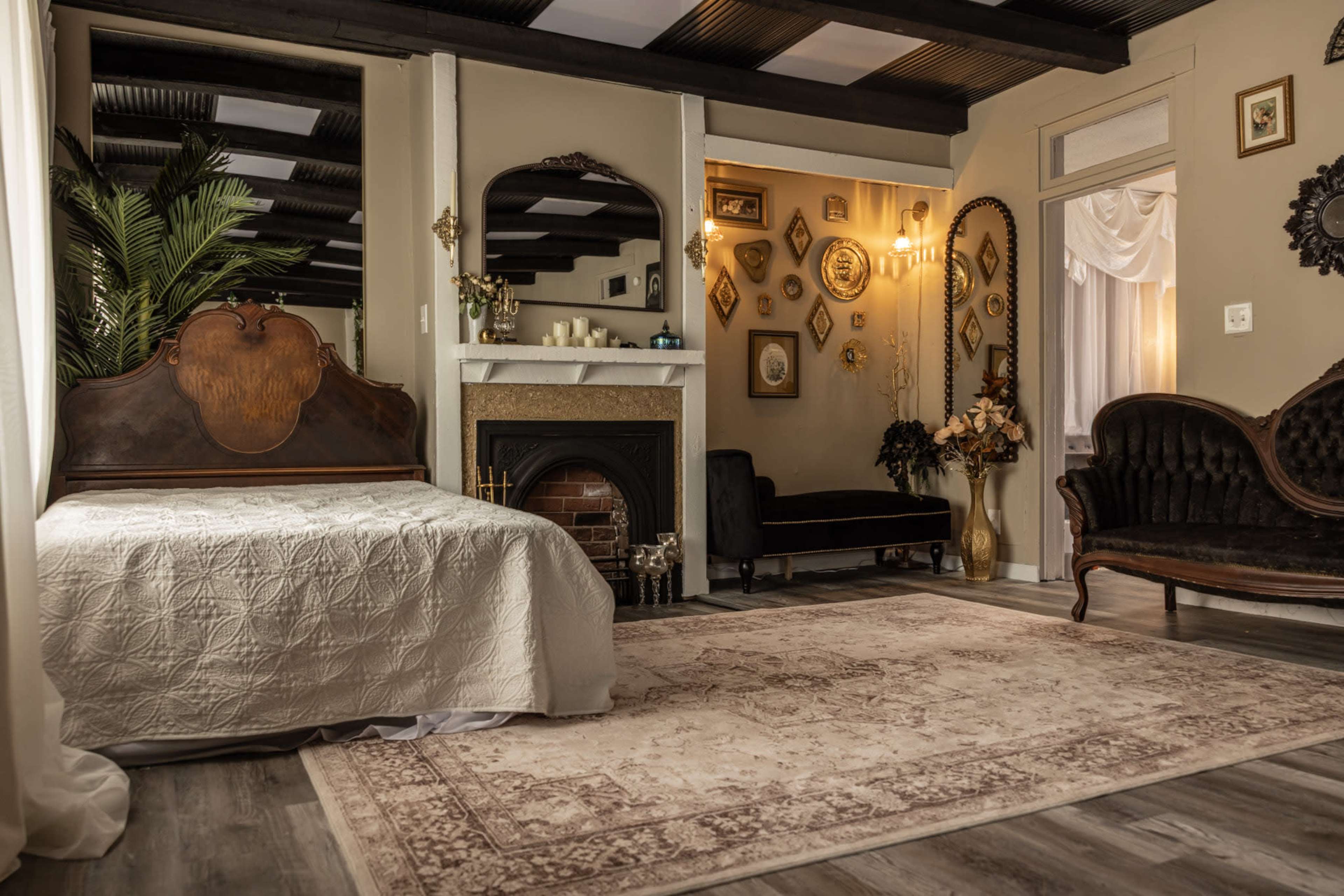 The image shows a cozy bedroom with a large ornate bed, a fireplace, a vintage-style seating area, and various decorative frames on the walls.