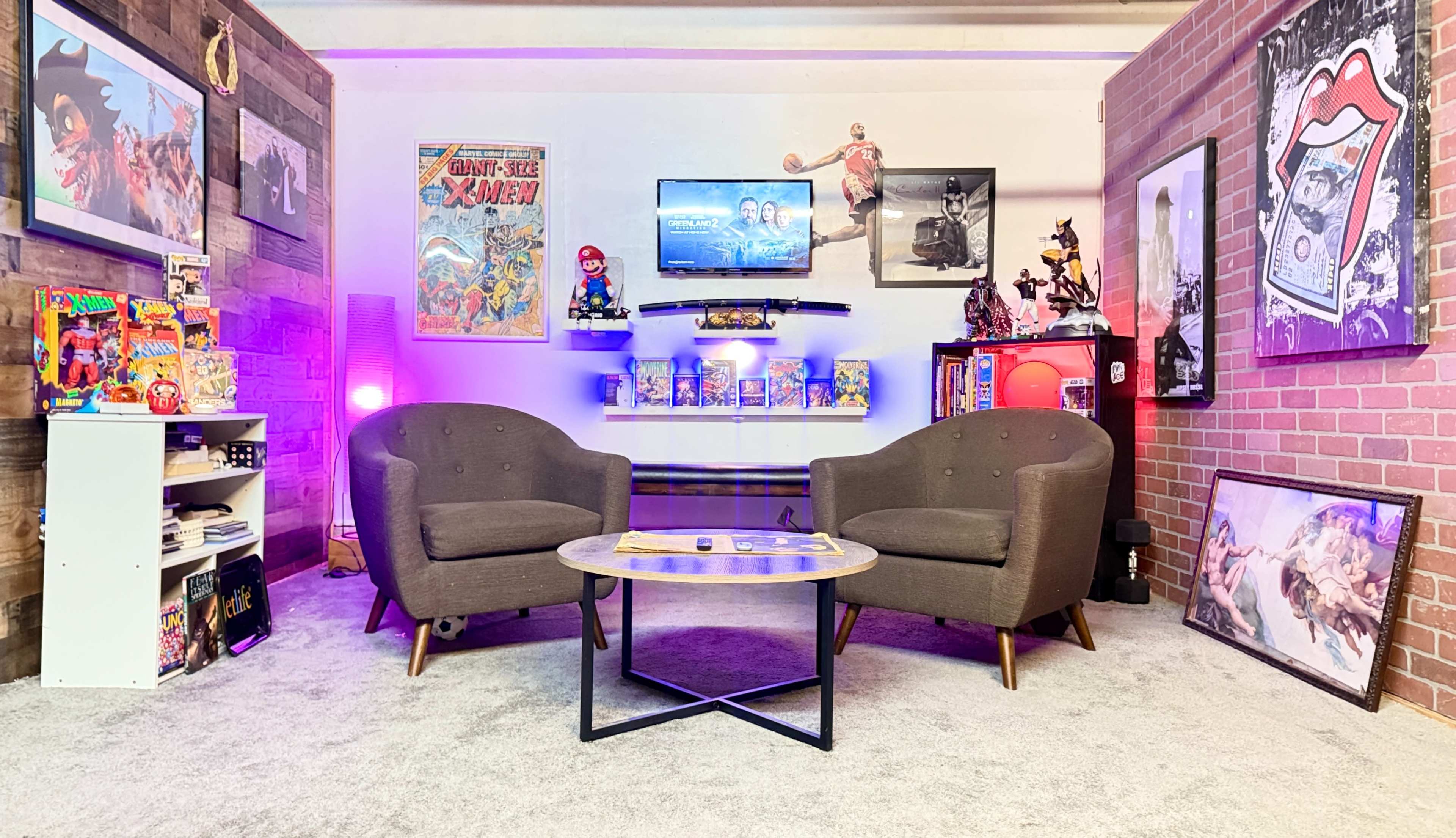 The image shows a cozy living room setup featuring two gray armchairs facing a coffee table, with colorful wall decor and shelves displaying various collectibles.