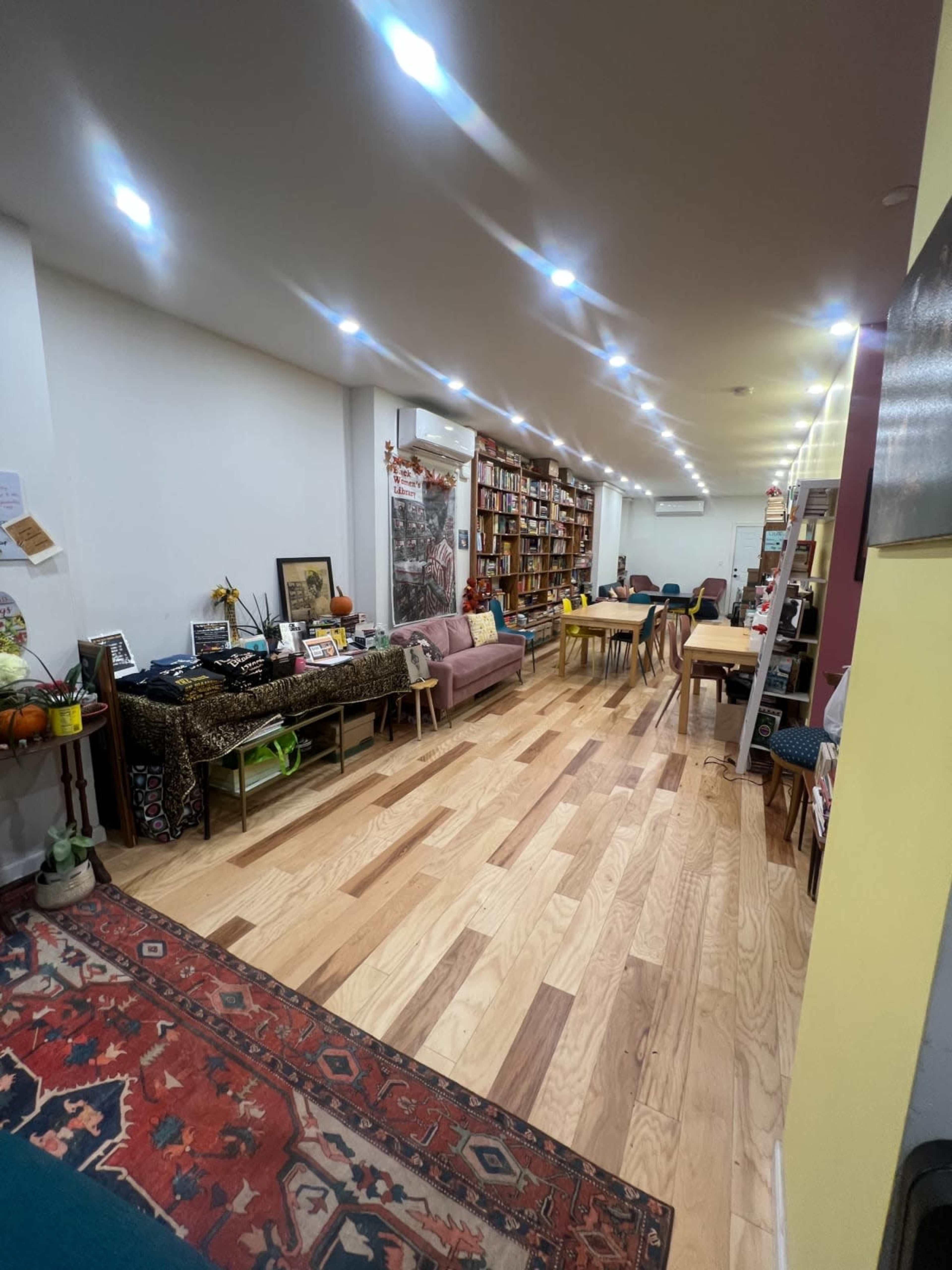 The image shows a spacious room with wooden floors, lined with bookshelves on one side, and various furniture including a sofa and tables scattered throughout.