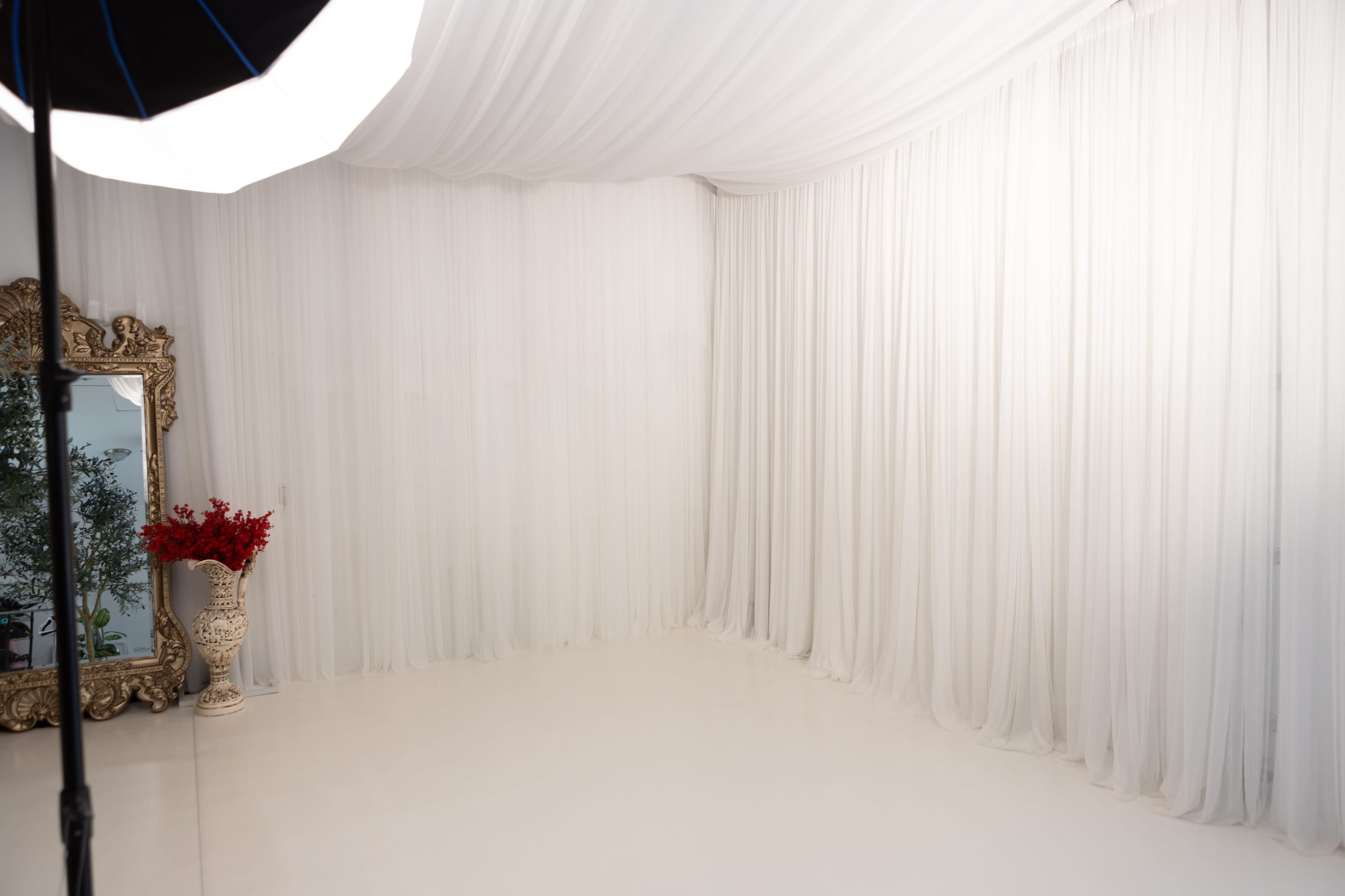 The image shows a bright, minimalist studio space with white draped fabric, a large ornate mirror, and a decorative vase with red flowers.