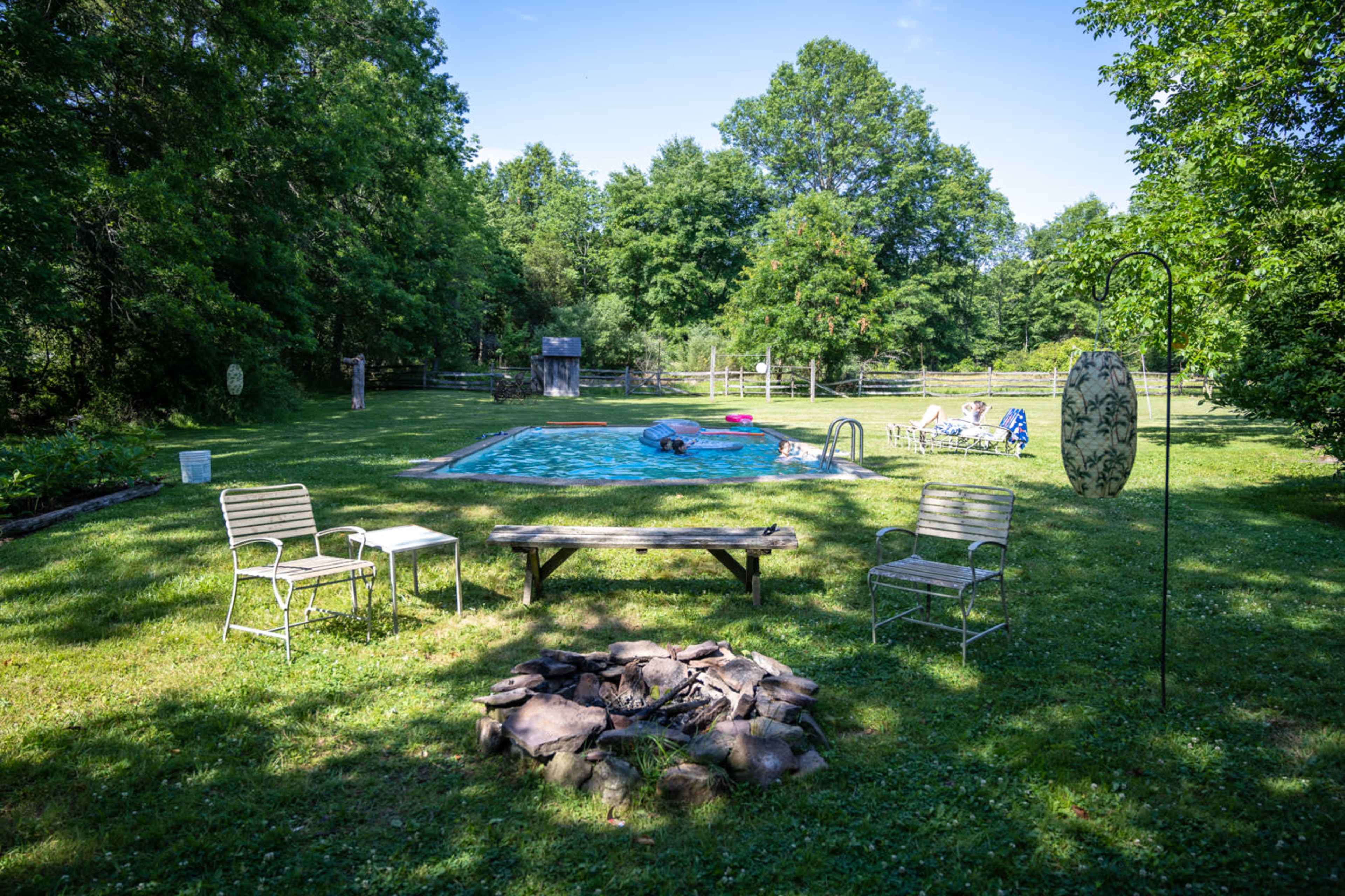 A swimming pool is surrounded by a grassy area with chairs, a fire pit, and trees in the background.