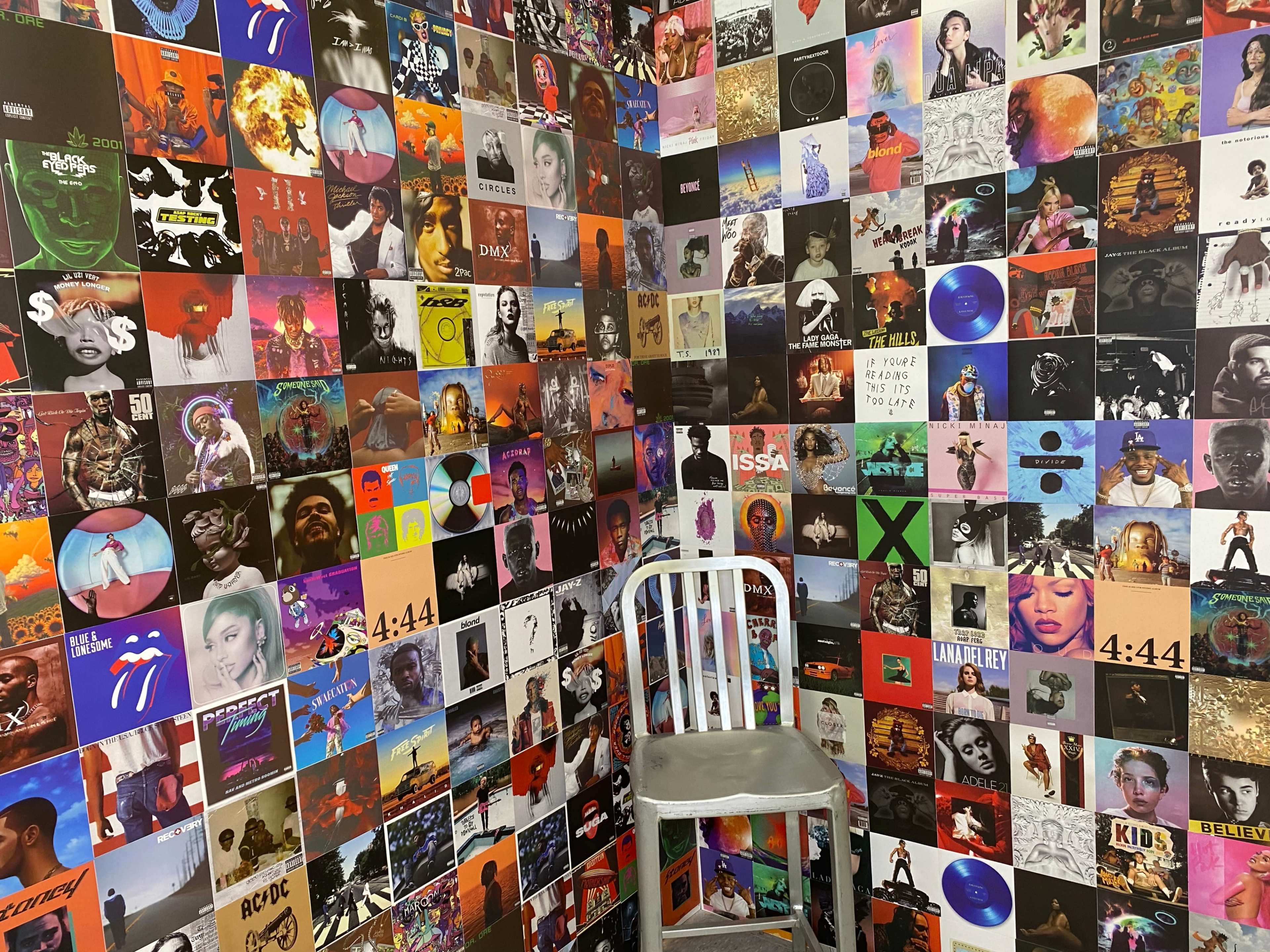 A corner of a room with walls covered in a collage of vibrant album covers, and a single white chair positioned in the corner.