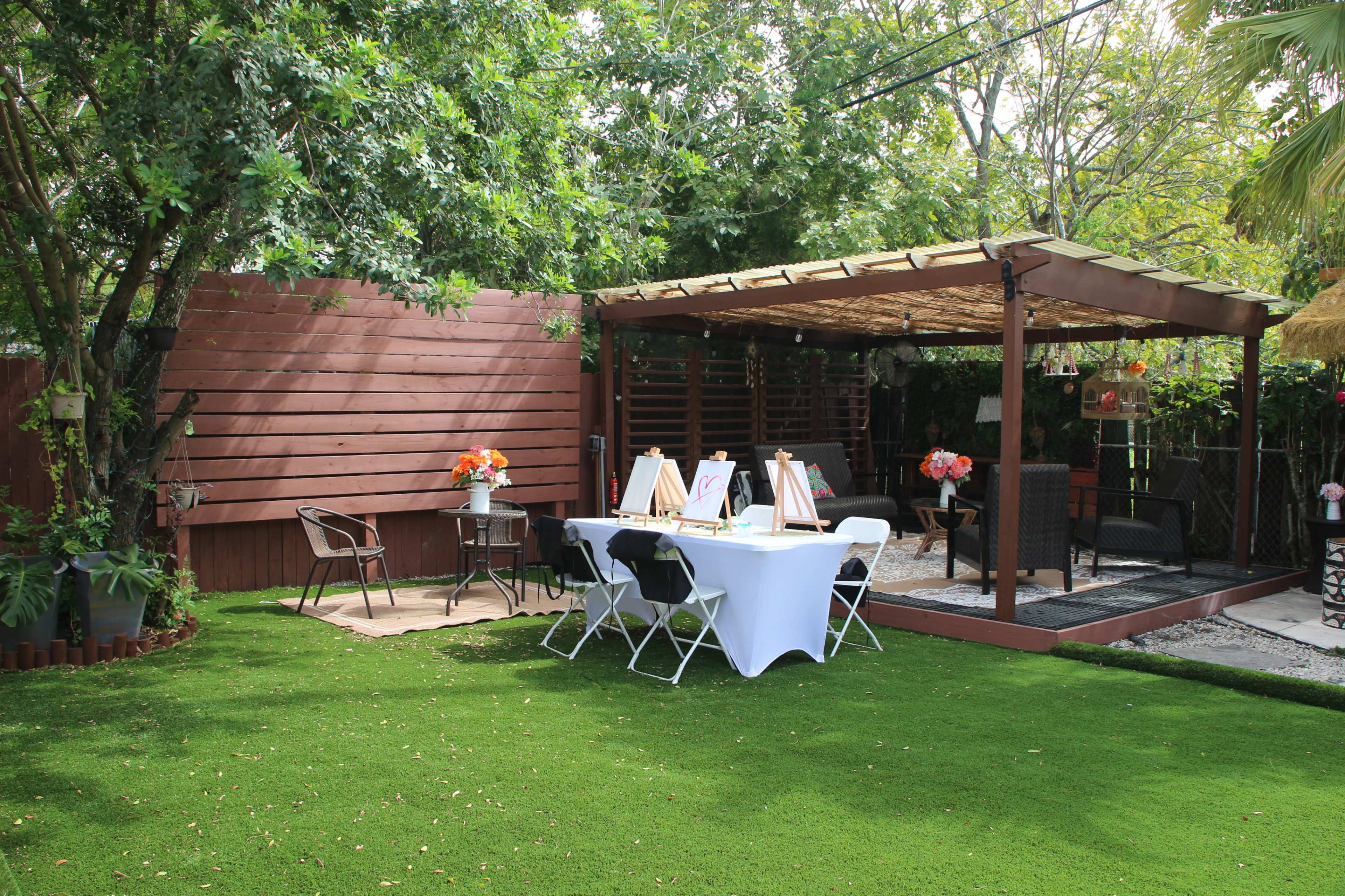 The image shows a landscaped backyard with a covered patio featuring seating and a table set up for painting, surrounded by greenery.
