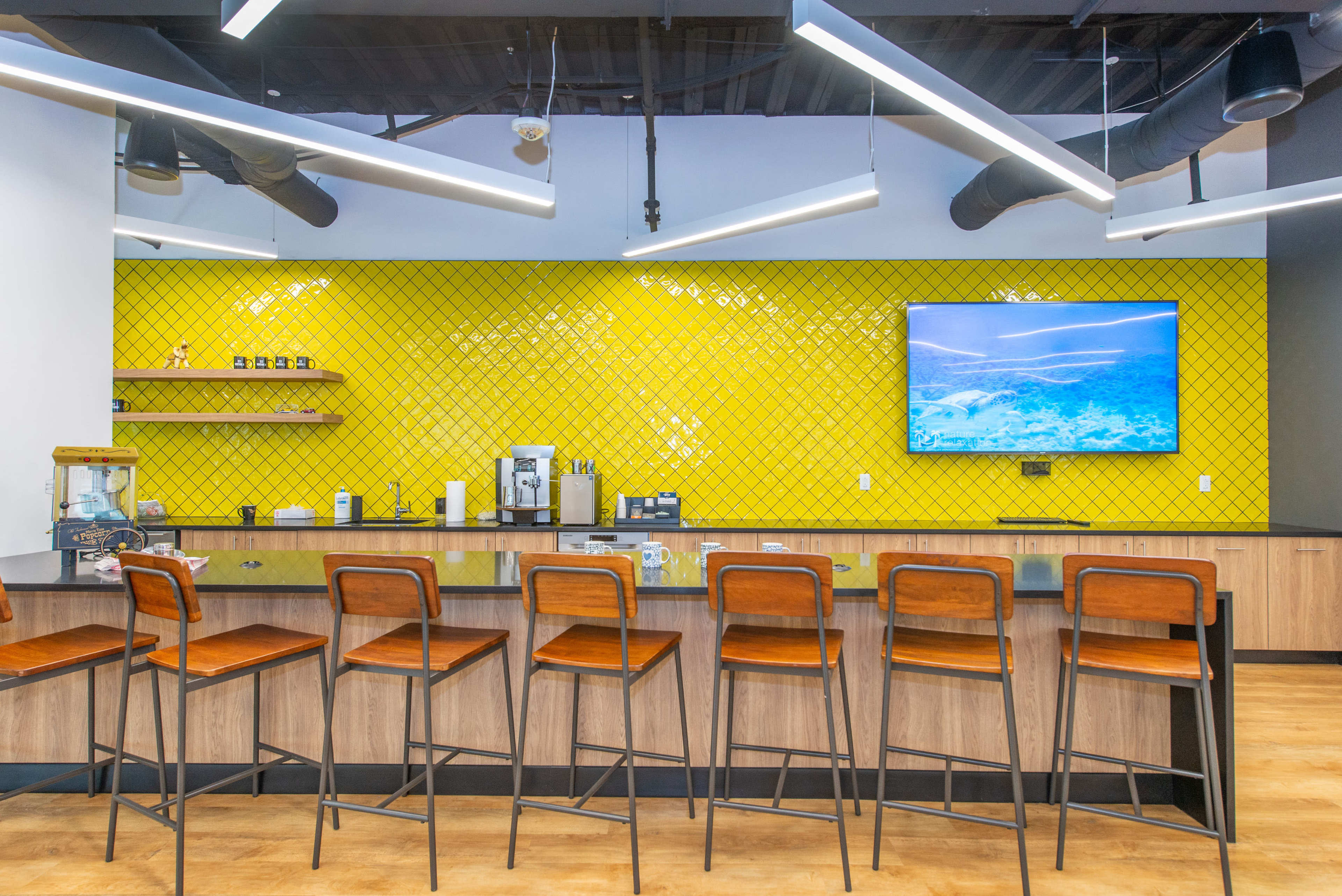 A modern kitchen area featuring a bar with high stools, a yellow patterned wall, and a television mounted above a countertop with various appliances.