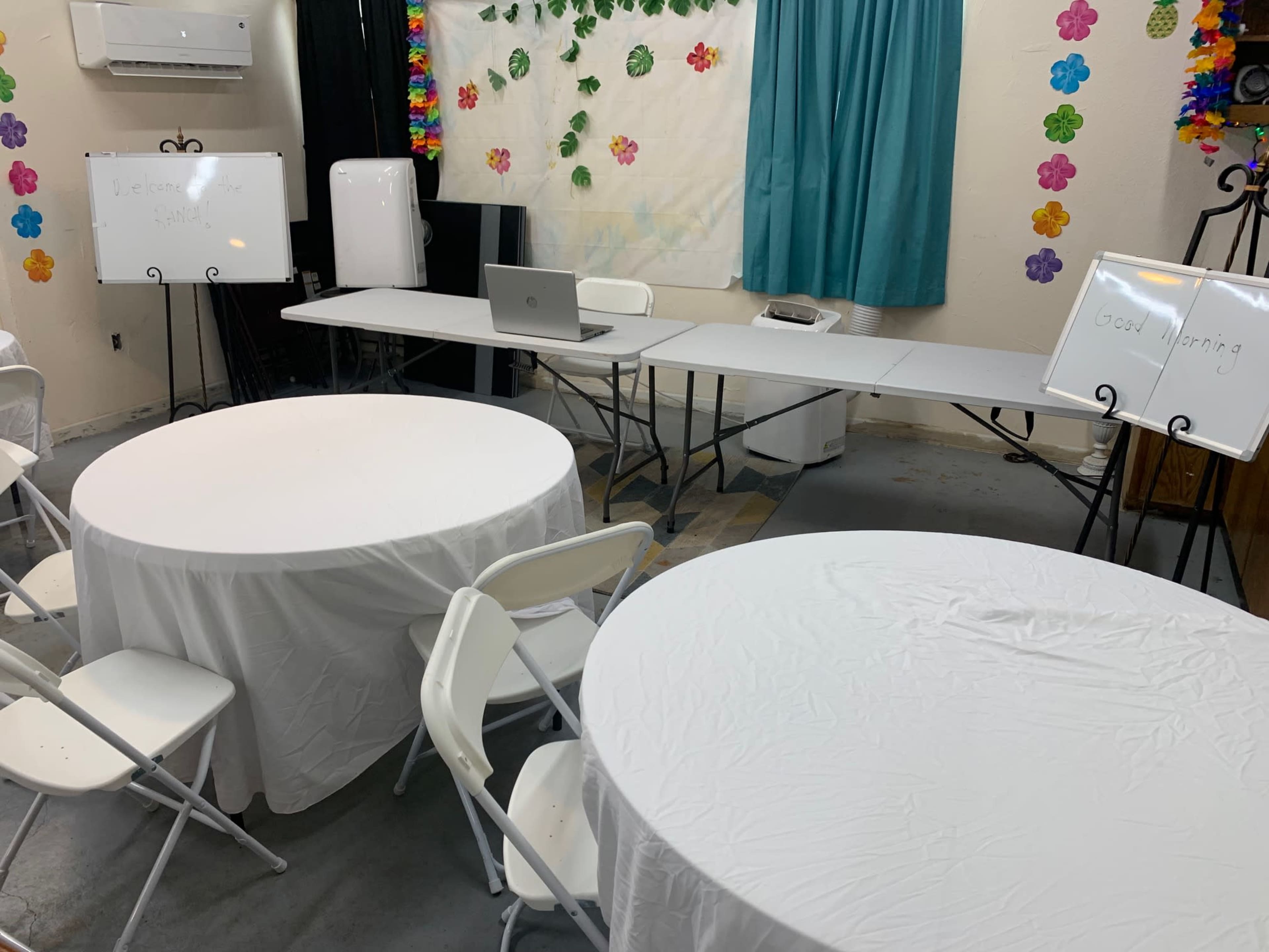 The image shows a room set up for a meeting or workshop, featuring two circular tables covered with white tablecloths, folding chairs, and a rectangular table with a laptop and other equipment.