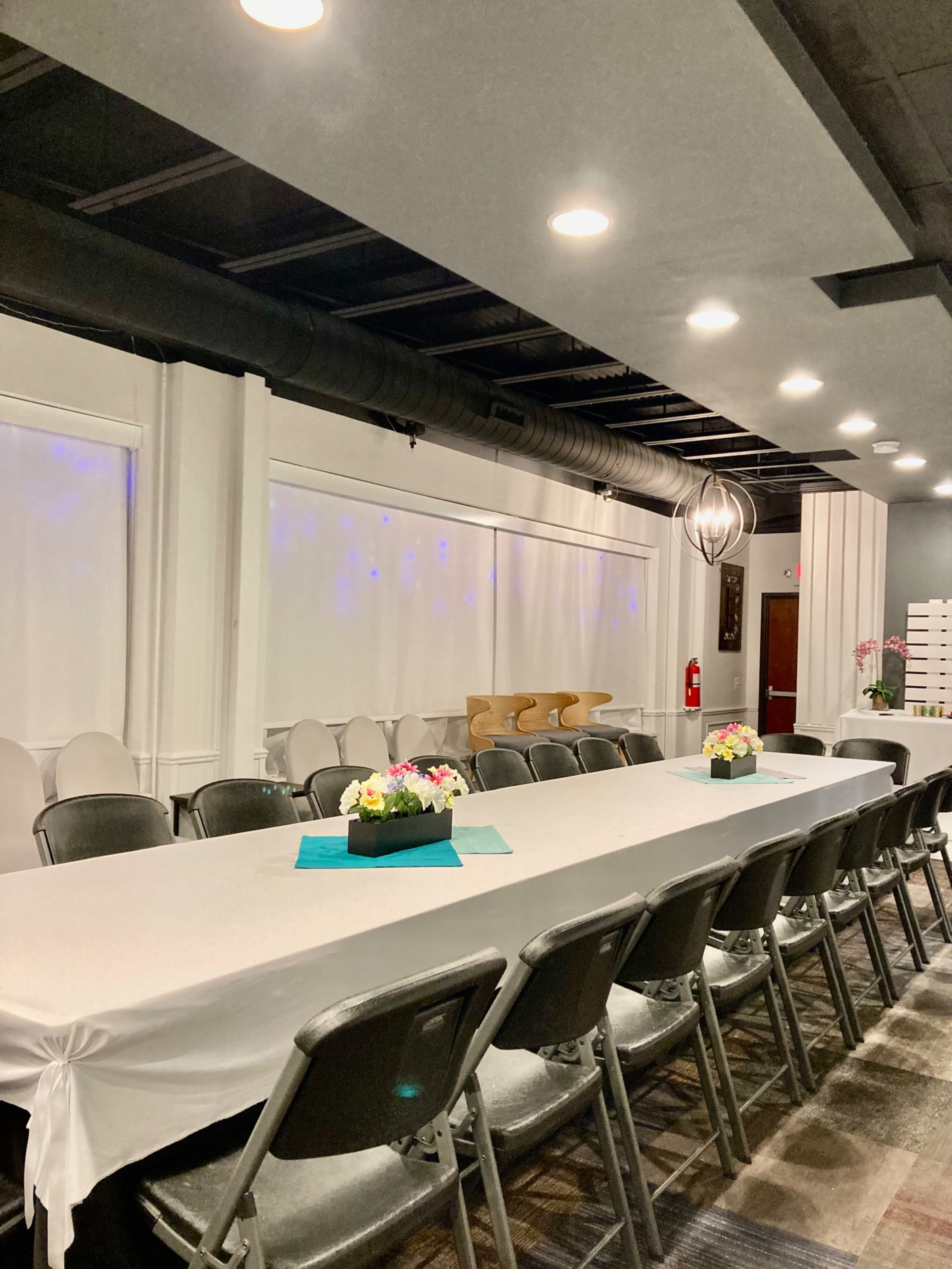 A long, white table with floral centerpieces is set in a spacious room featuring overhead lighting and gray walls.