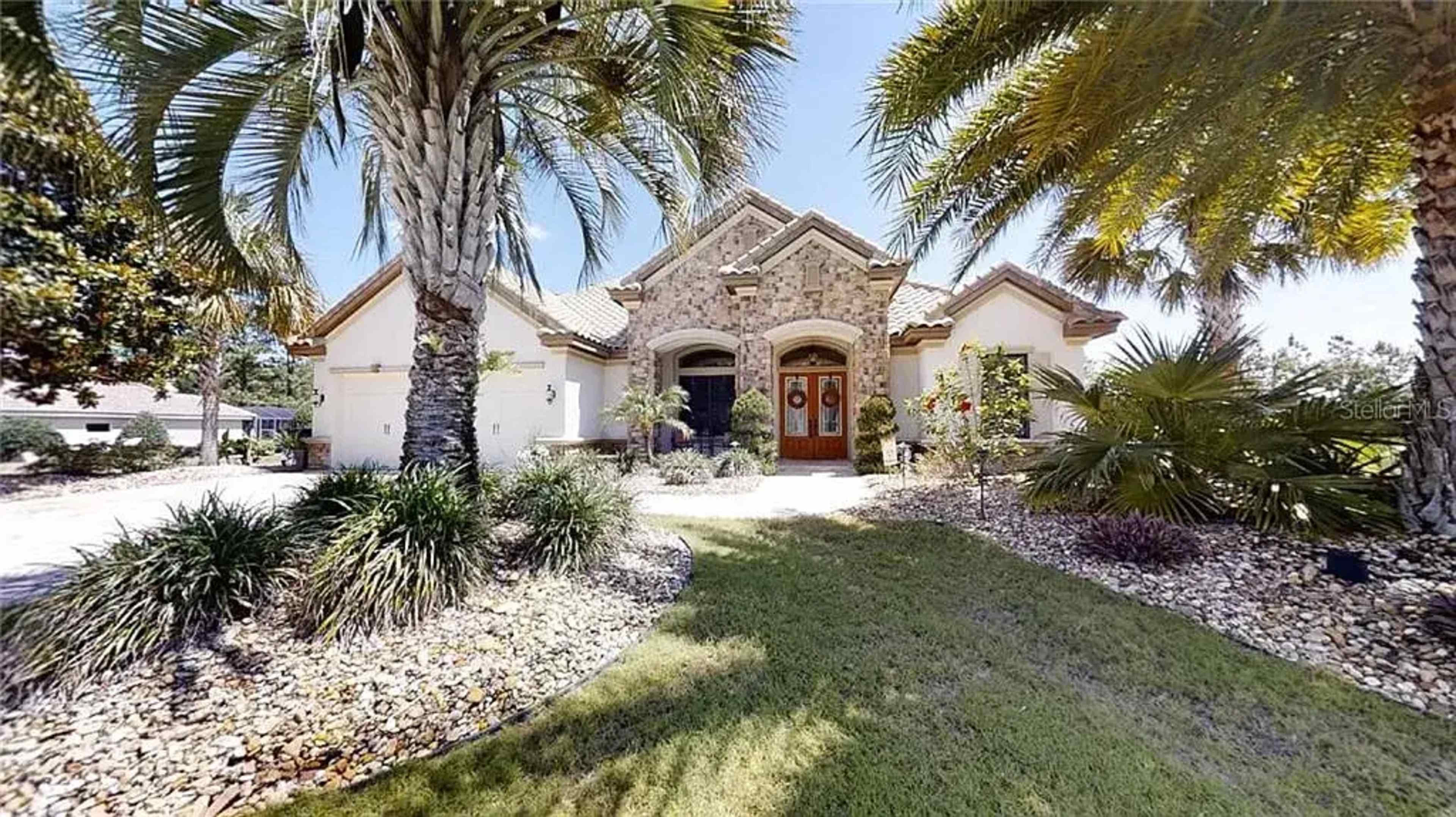 A large stone-front house with a tiled roof is framed by palm trees and landscaped greenery in the front yard.