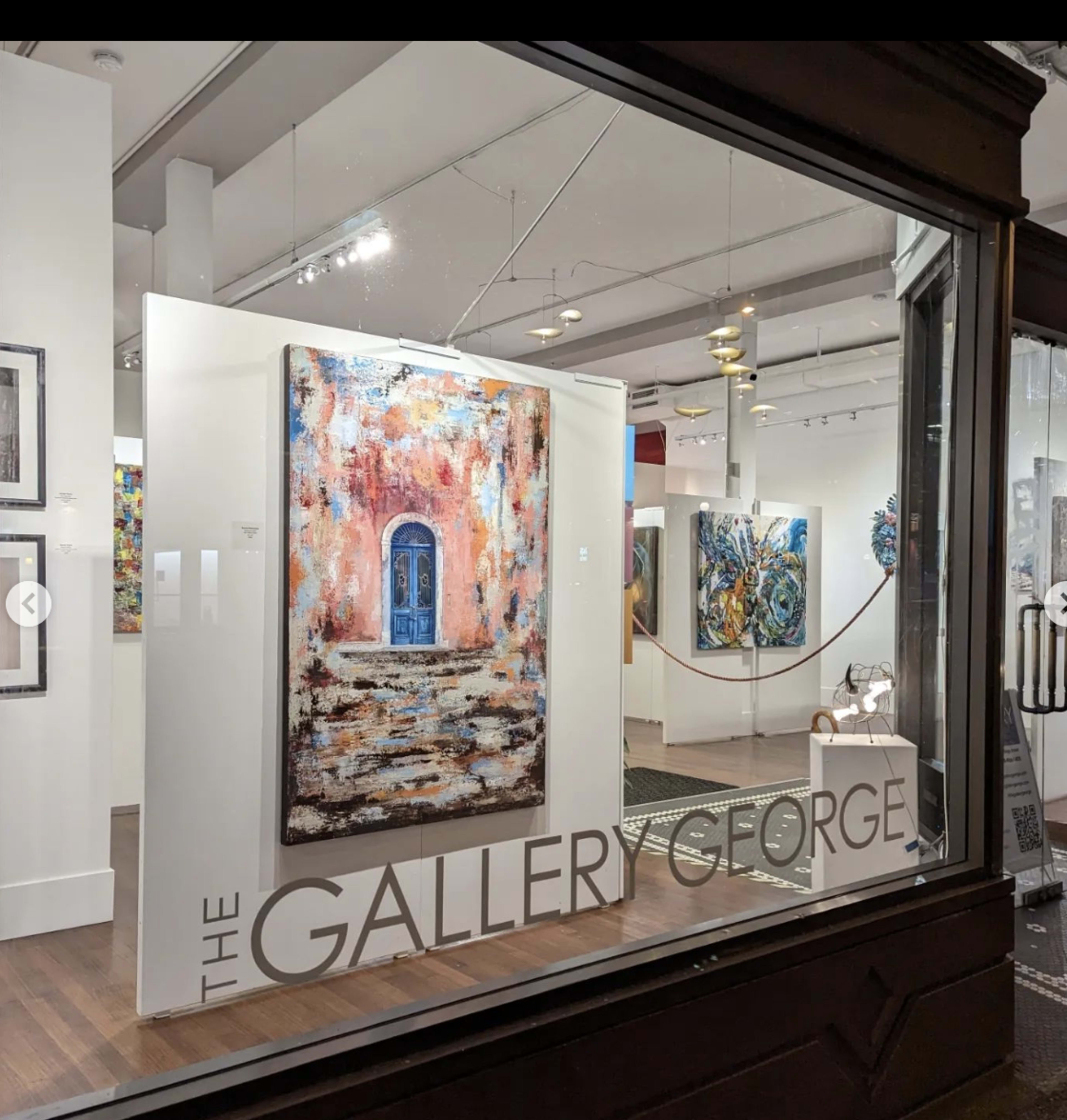 A view of an art gallery's interior, featuring a large painting of a blue door on a pink wall displayed prominently behind a glass window.