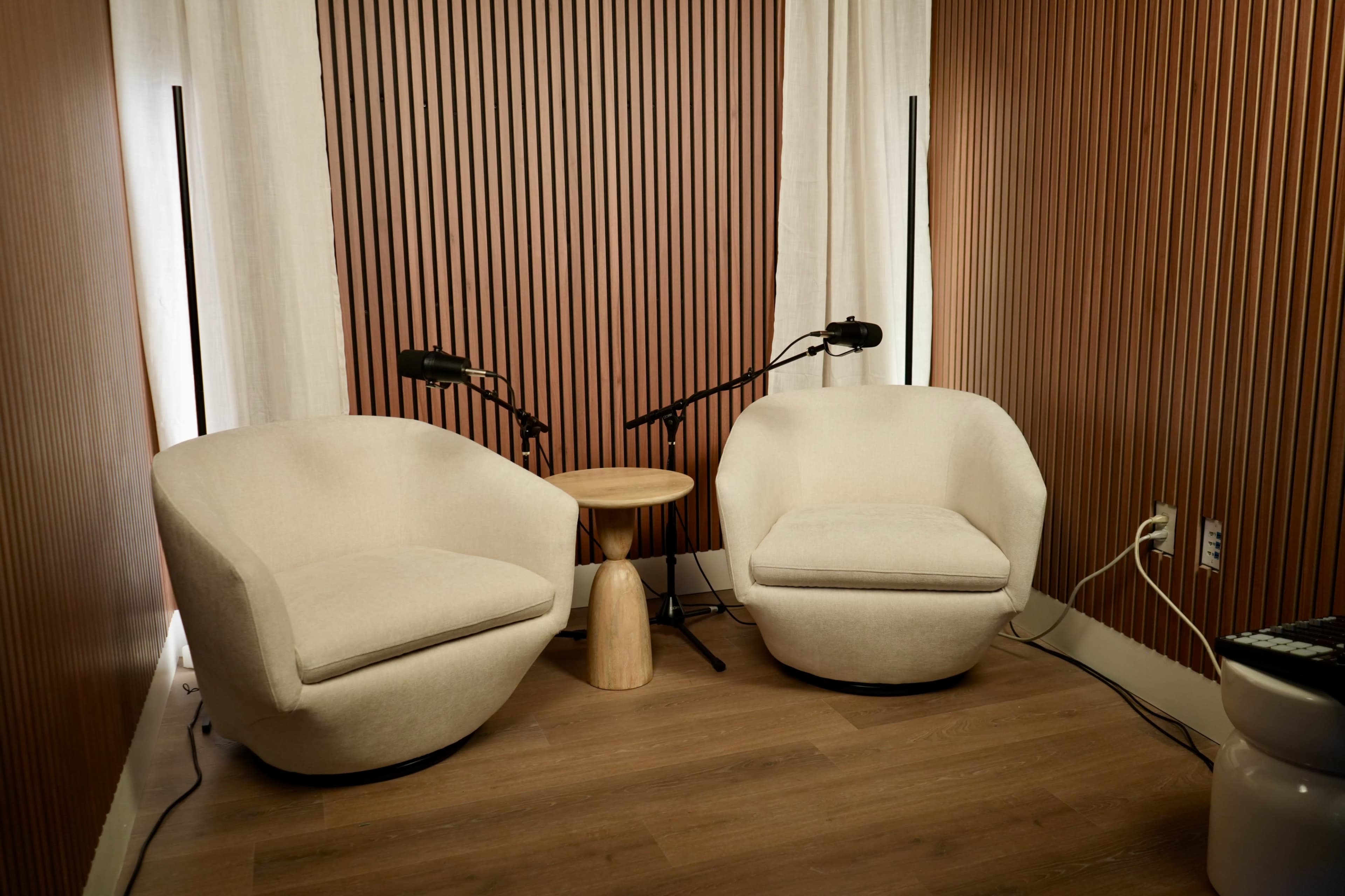 The image shows a small, cozy sitting area featuring two round, beige chairs positioned around a wooden side table, with two microphones set up nearby.