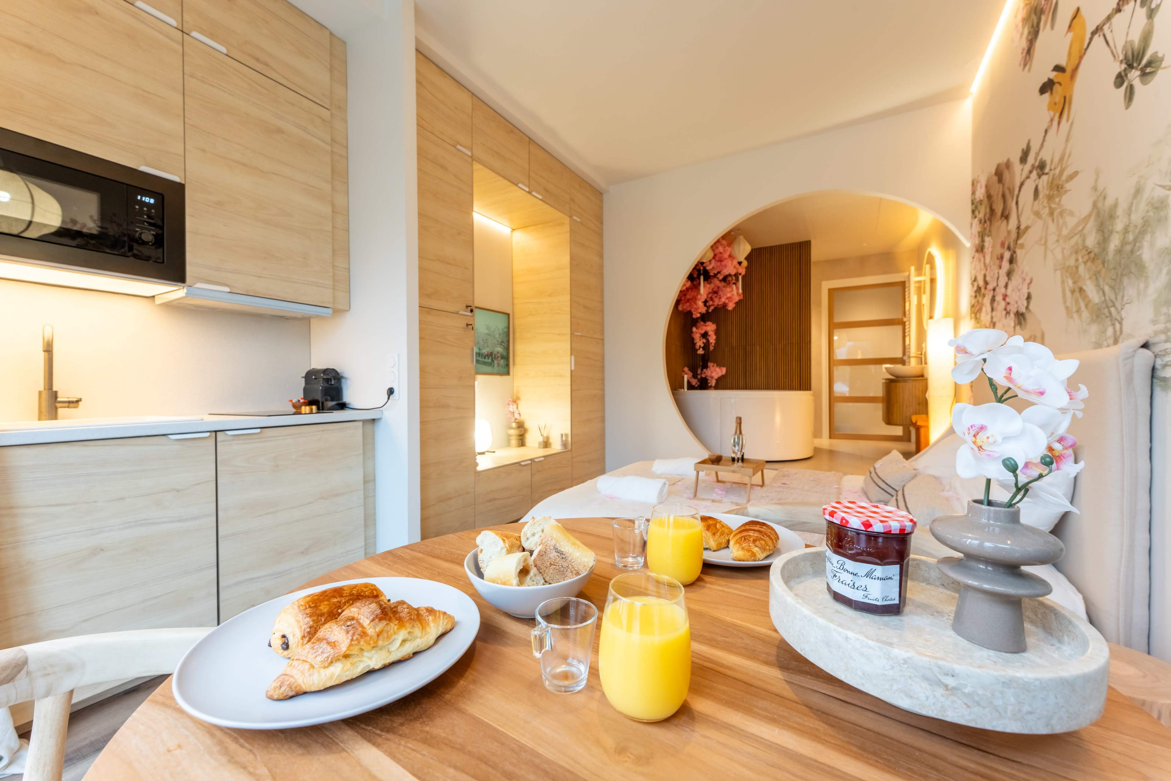 A wooden table is set with pastries, orange juice, and a decorative vase, while a modern kitchen and softly lit hallway are visible in the background.