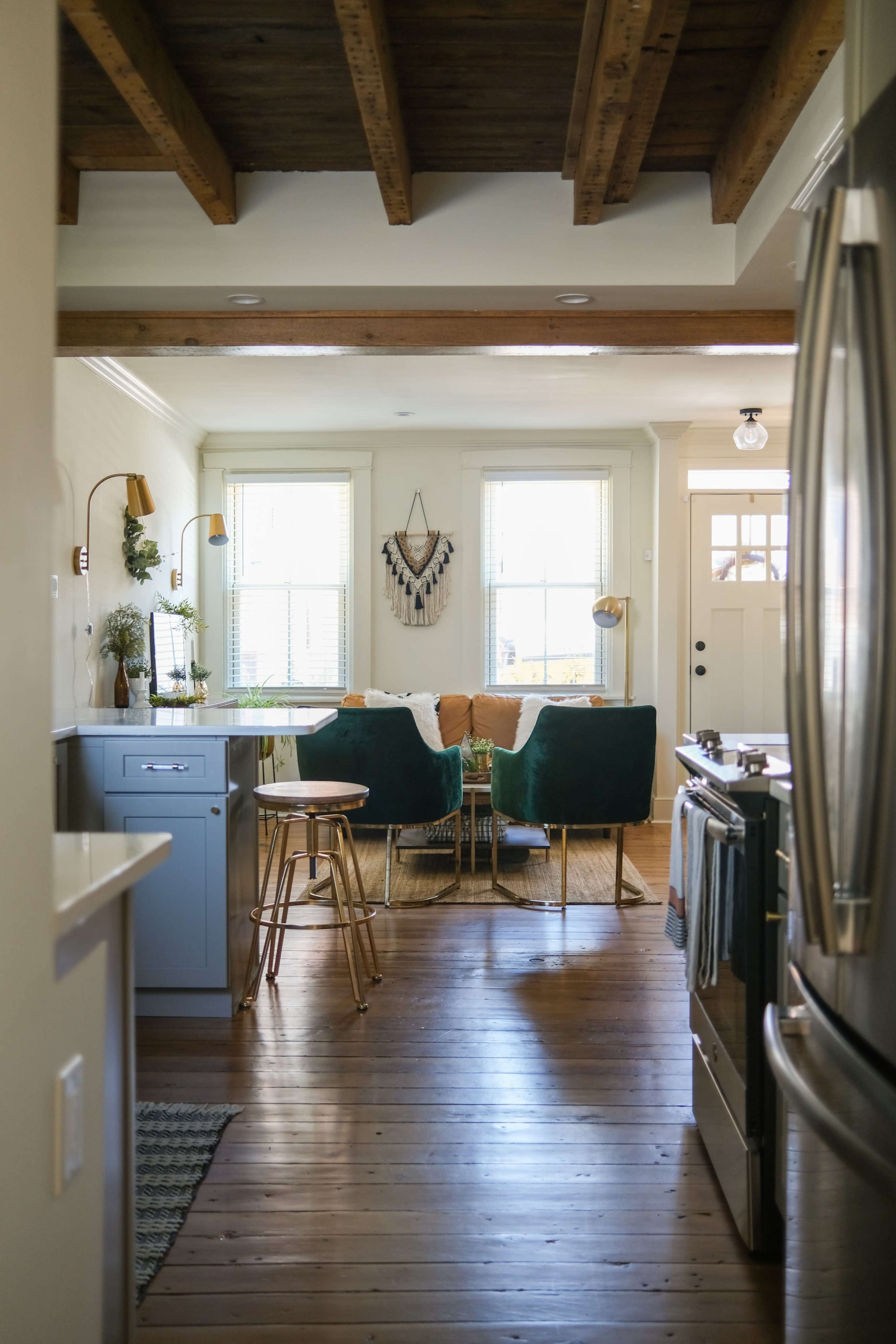 A well-lit kitchen opens into a living area decorated with green chairs and a wooden table, featuring large windows and exposed beams above.