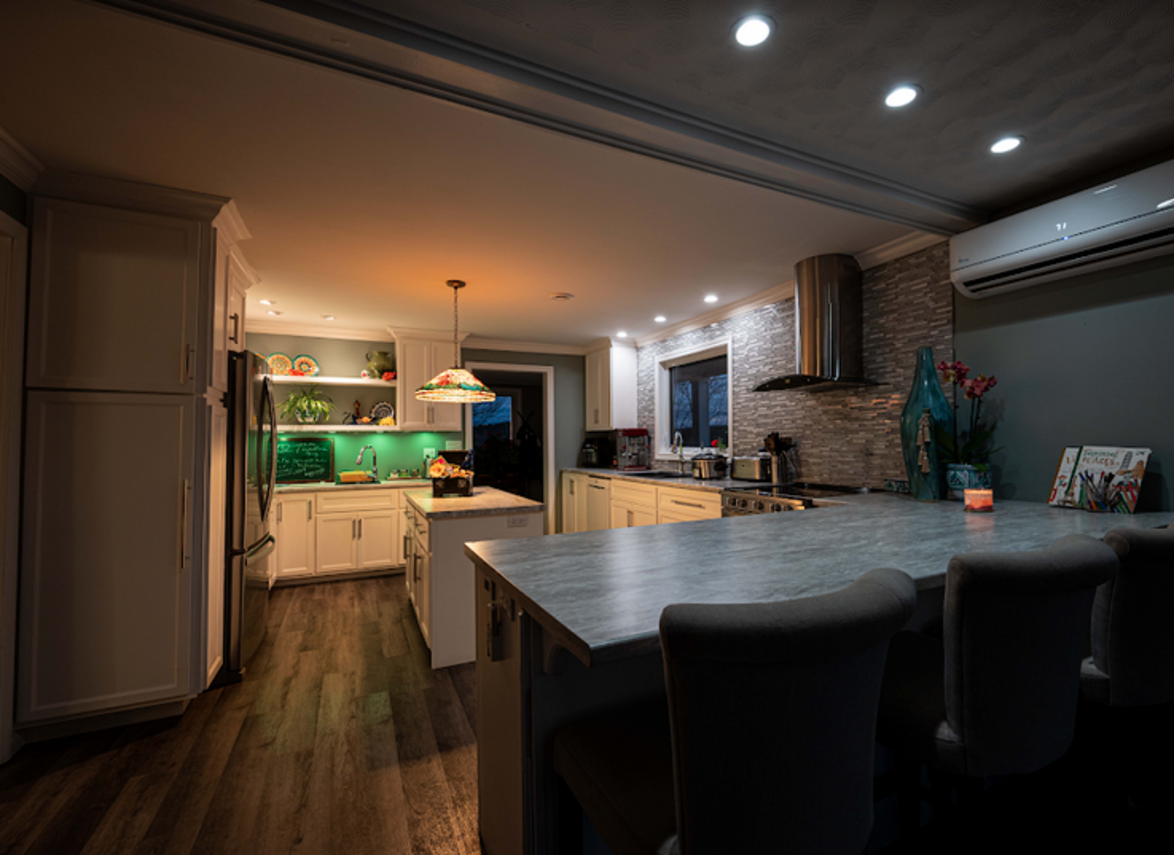 A modern kitchen features white cabinetry, a gray countertop, pendant lighting, and a green accent wall illuminated by under-cabinet lights.