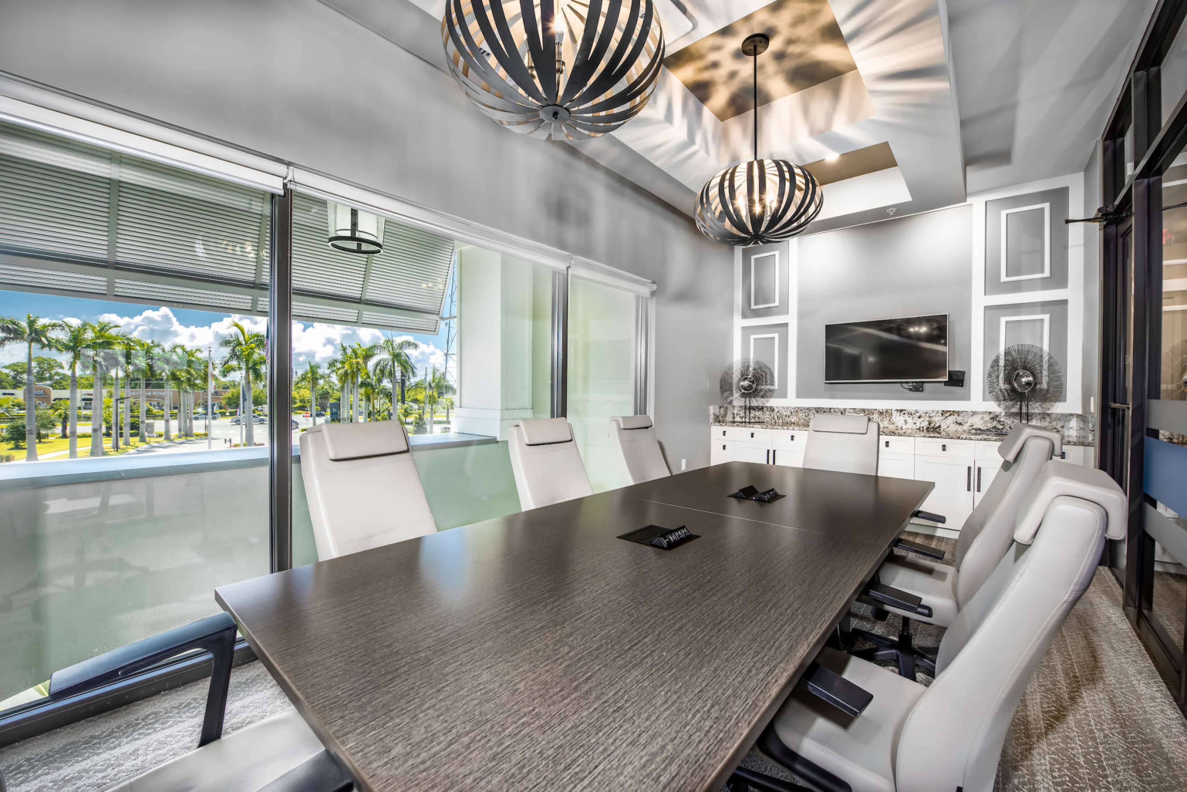 A modern conference room featuring a long wooden table, white leather chairs, and large windows with a view of palm trees outside.