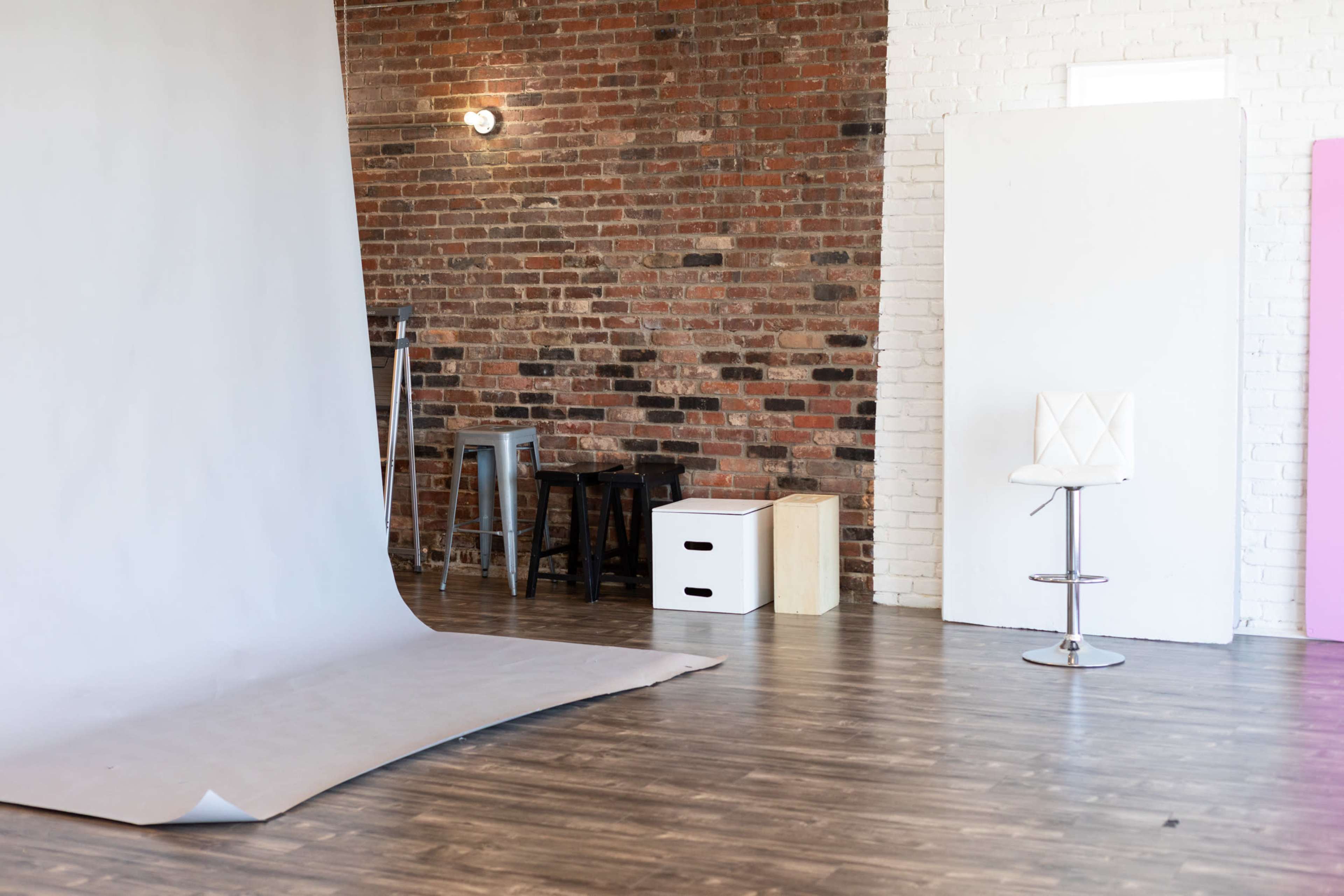 The image shows a photo studio with a gray backdrop, a white stool, and various studio props against a brick and white wall.