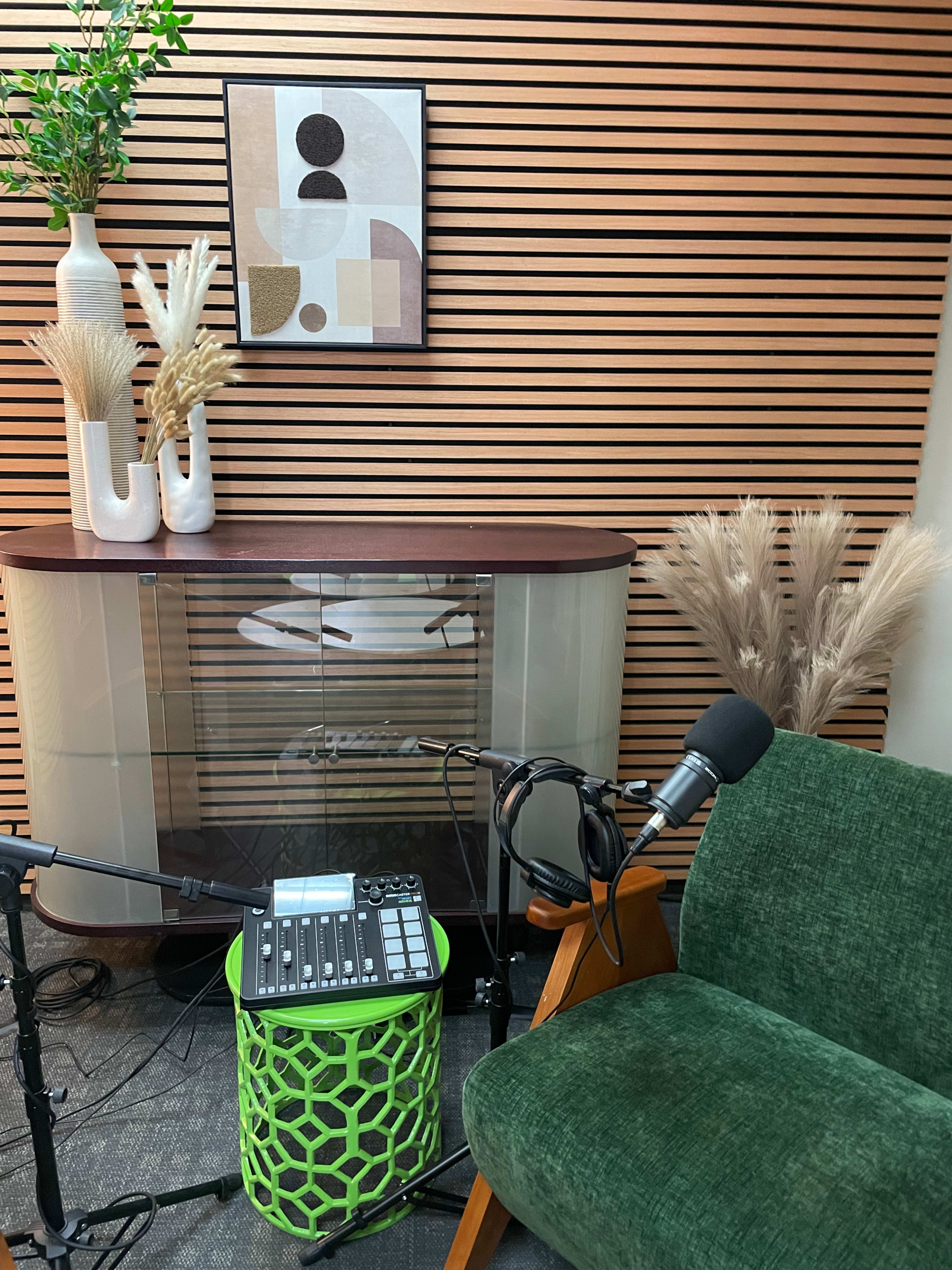 The image shows a modern recording setup featuring a green chair, a microphone, and a table with decorative items against a backdrop of wooden paneling.
